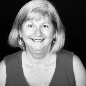 A black-and-white portrait of a smiling middle-aged woman with shoulder-length hair, wearing earrings and a sleeveless top.