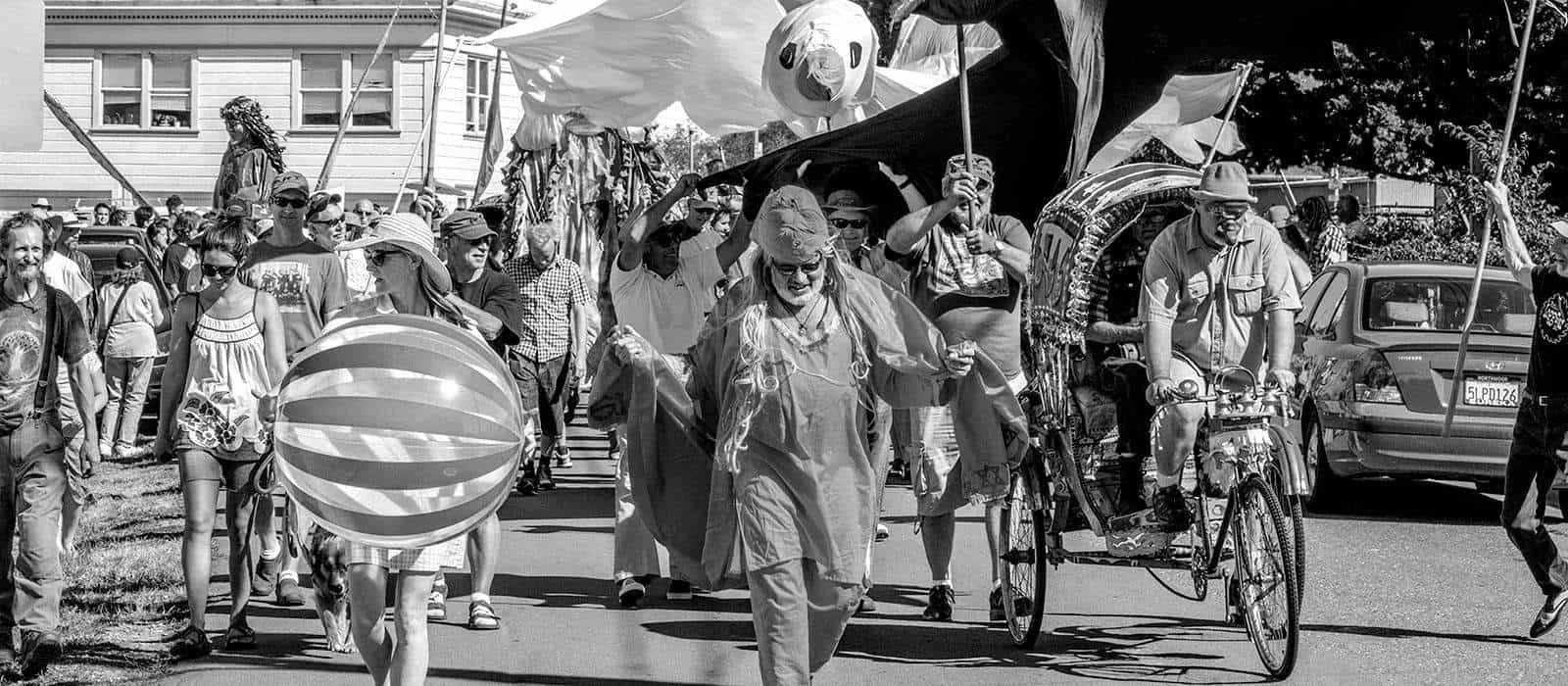 Crowd of people walking on the street during a parade with some individuals riding decorated bicycles, carrying balloons, and dressed festively.