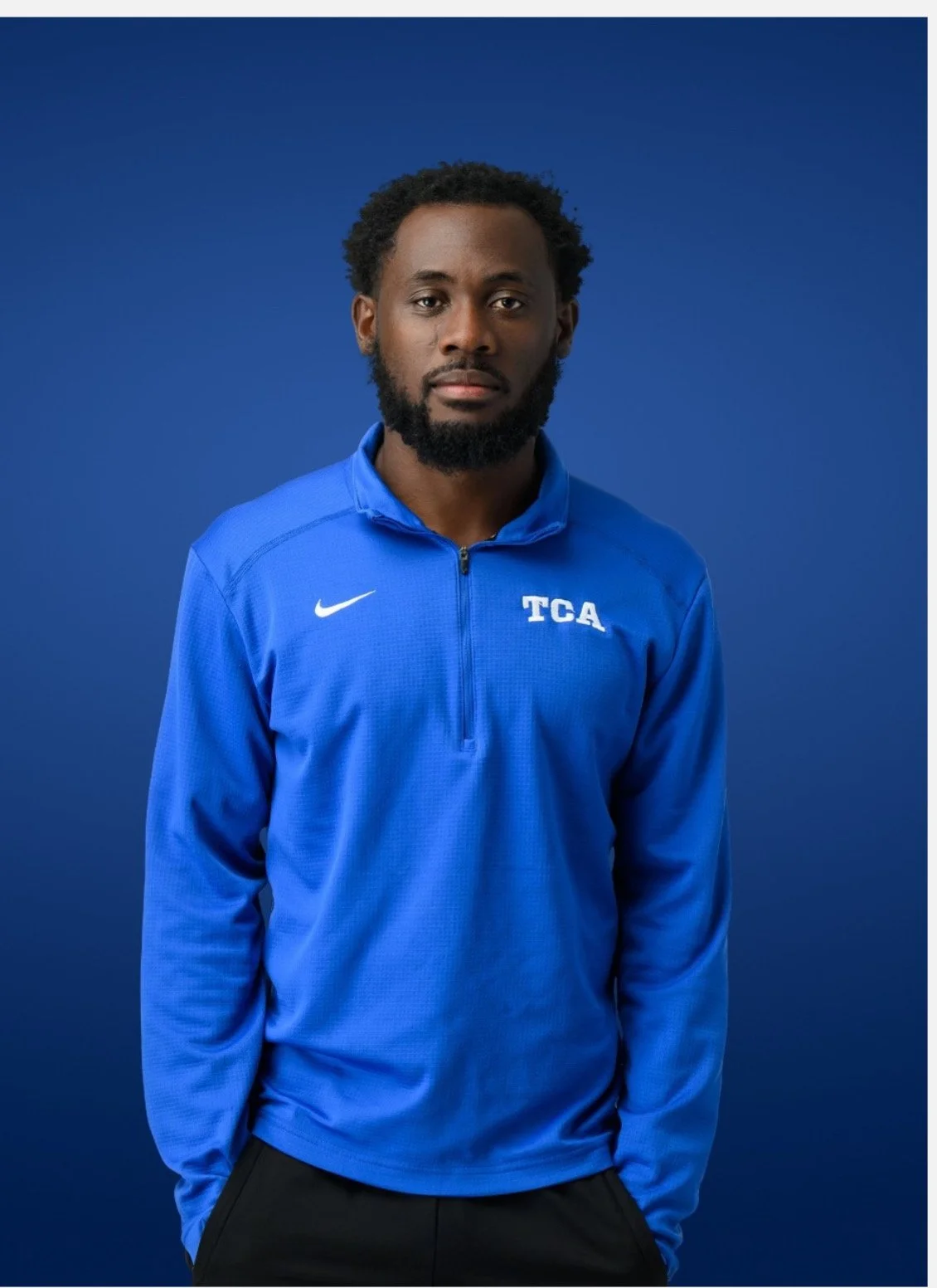 Portrait of a man with a beard wearing a blue athletic jacket with a Nike logo and 'TCA' initials, standing against a blue background.