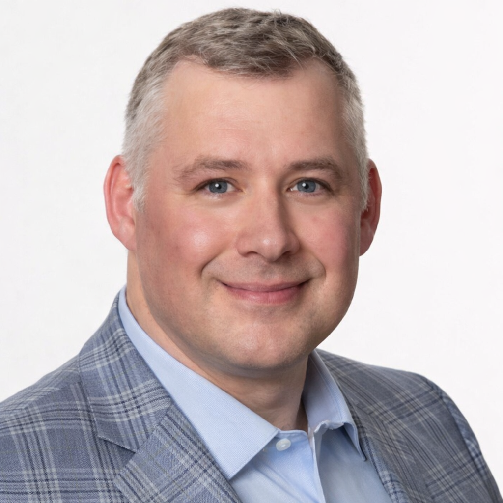 Professional headshot of a man with short, light-colored hair, wearing a light blue collared shirt and a gray plaid blazer, smiling against a plain white background.