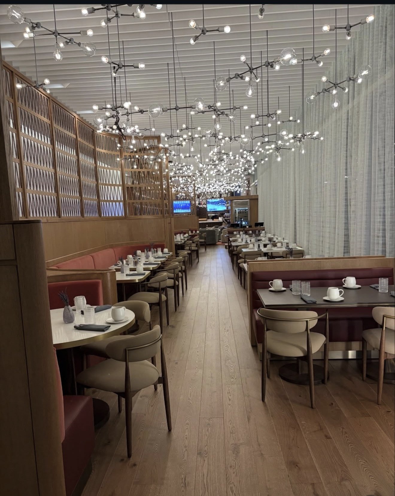 Empty restaurant dining area with wood flooring, tables set for dining with cups and glasses, beige and maroon chairs, decorative string lights on ceiling, and wooden divider on the left side.