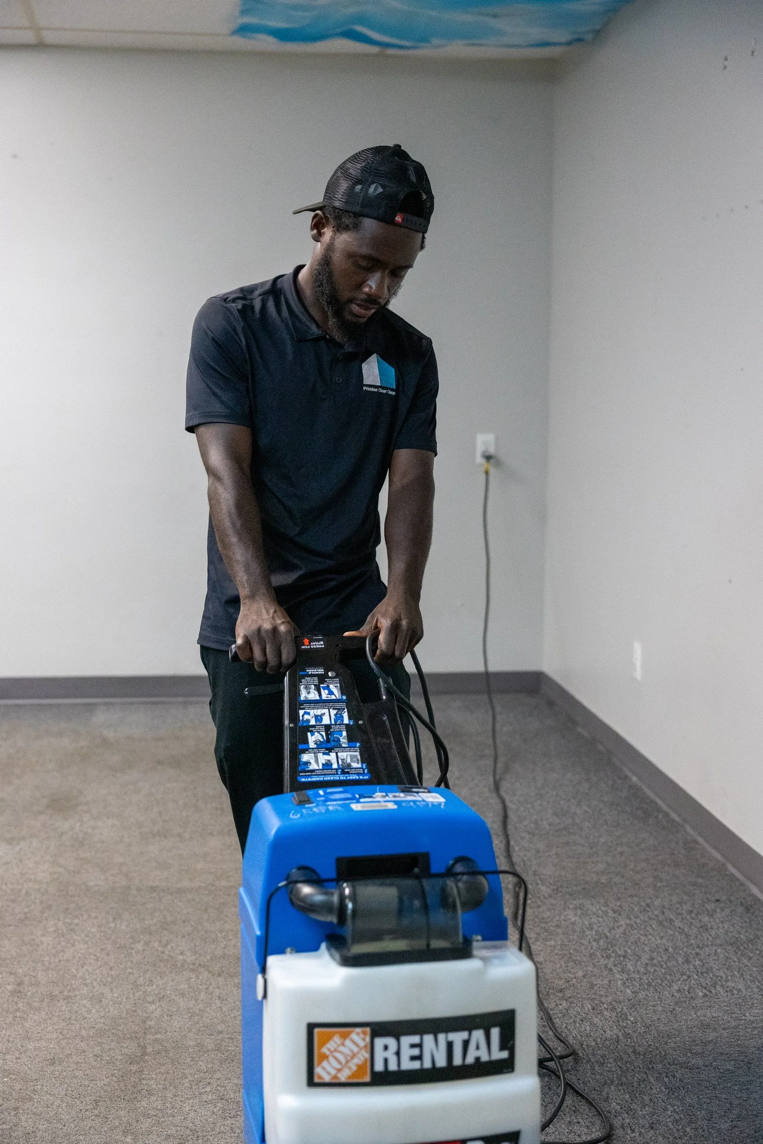 A man operating a carpet cleaning machine in a room with plain walls and carpeted floor.