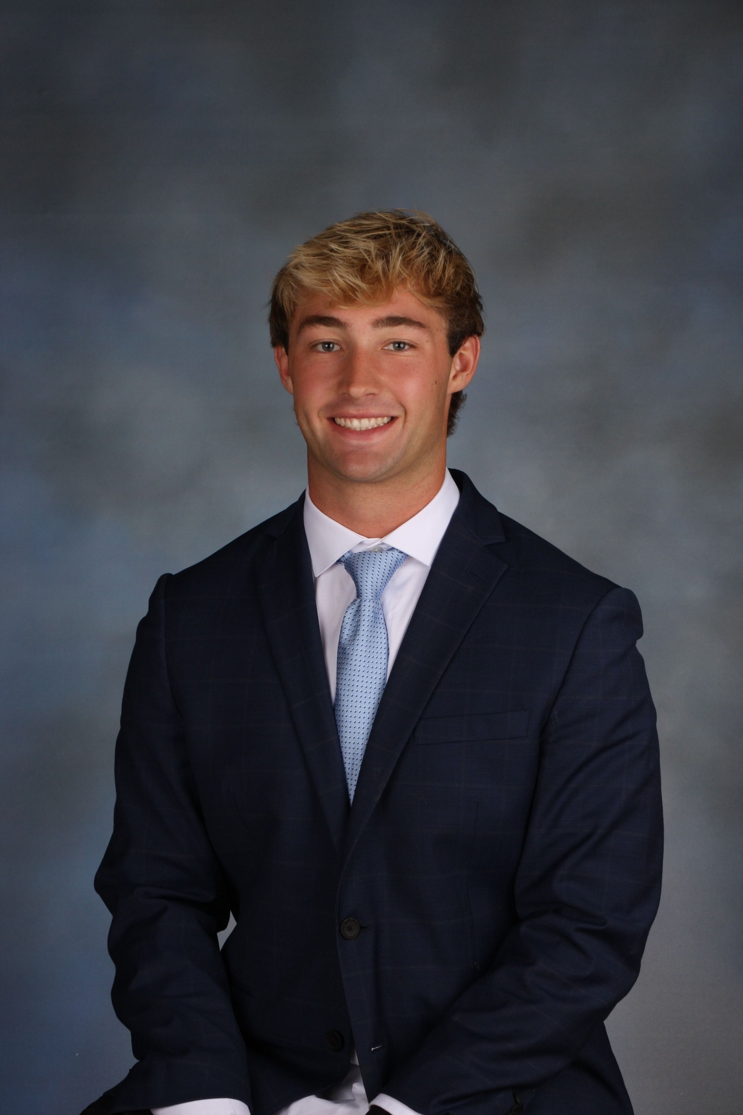 A young man in a dark navy suit, white shirt, and light blue tie, smiling, posed for a formal portrait against a cloudy gray background.