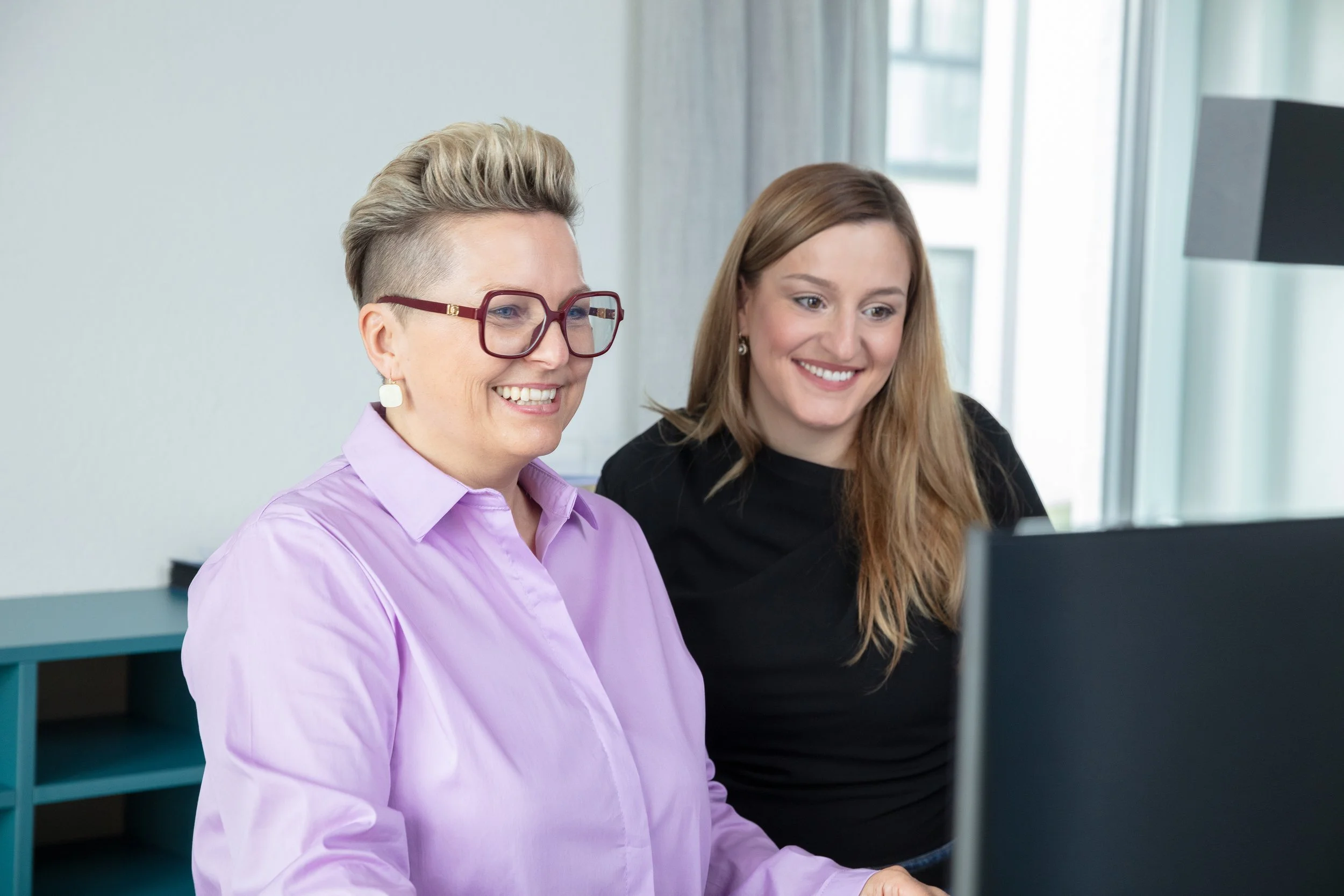 Regina Lackner im Gespräch mit einer jungen Kollegin. Sie erklärt ihr etwas am Computer. Ausbildung und Nachwuchsförderung.