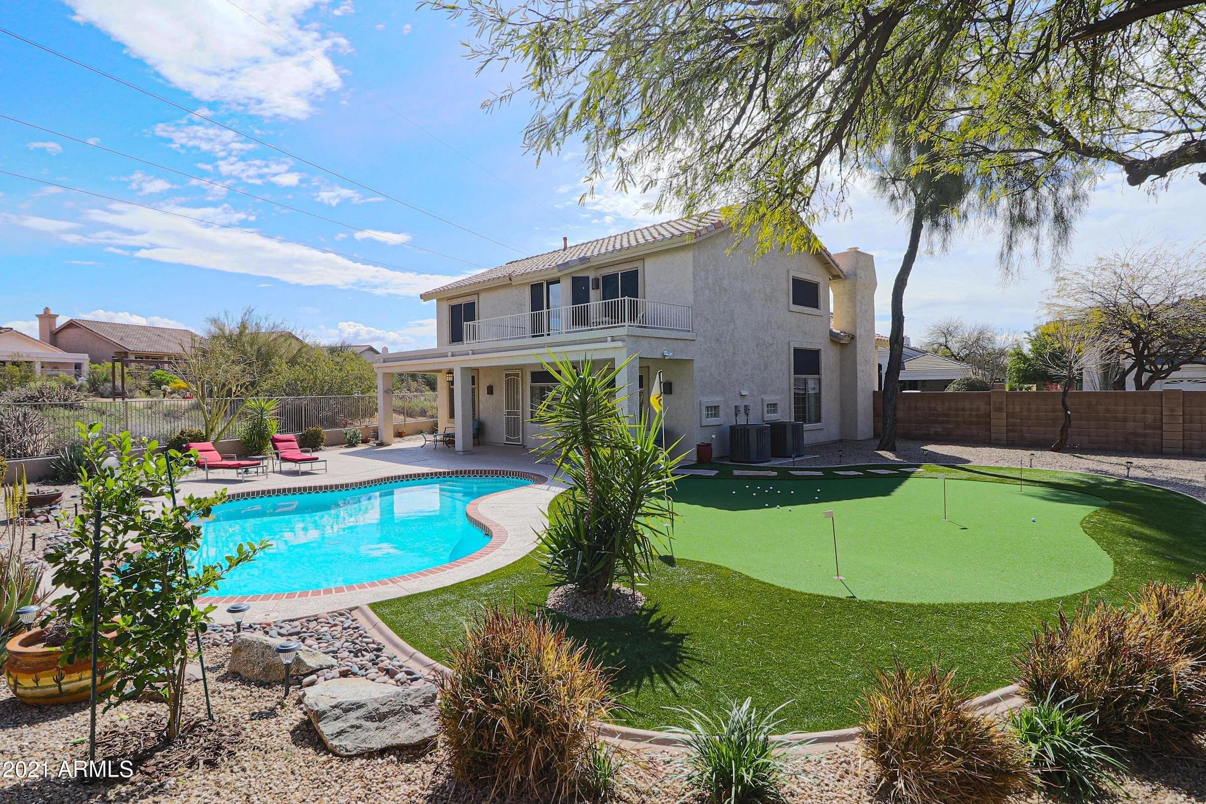 Backyard with a swimming pool, green artificial turf putting green, lounge chairs, and a two-story house with a balcony under a blue sky with scattered clouds.