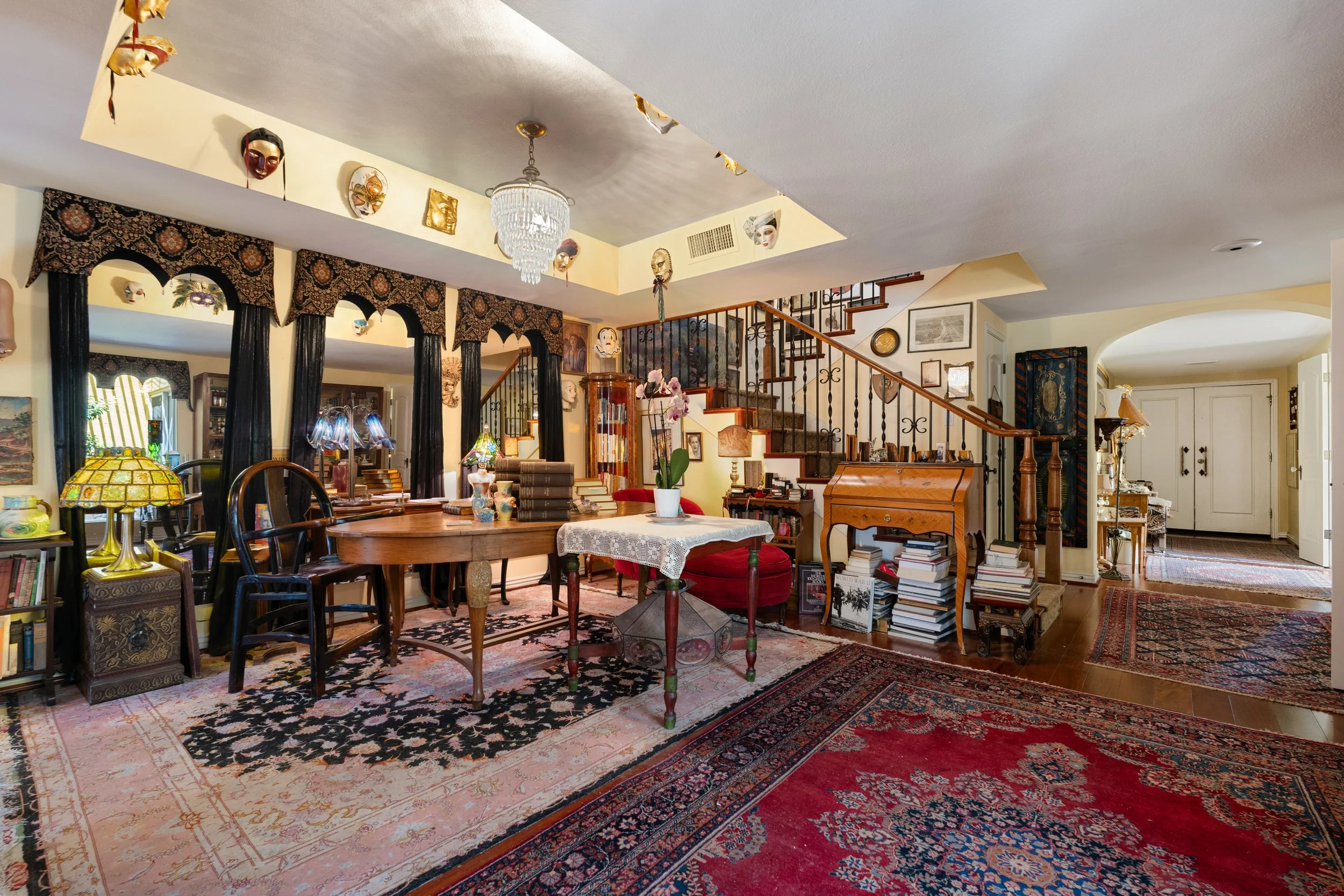 A cozy library room with a high ceiling decorated with various masks. The room features a large, dark patterned valance with black curtains, a round wooden table with books, a stained glass lamp, and a floral arrangement. Stairs lead to a second floor, and the room has multiple framed pictures and decorative items, with Persian-style rugs on the wooden floor.
