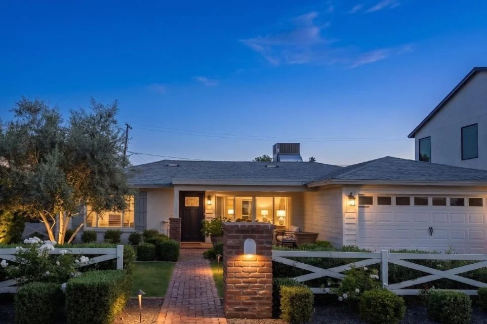 A cozy suburban house at dusk with a brick pathway, lush greenery, a white fence, and exterior lighting.