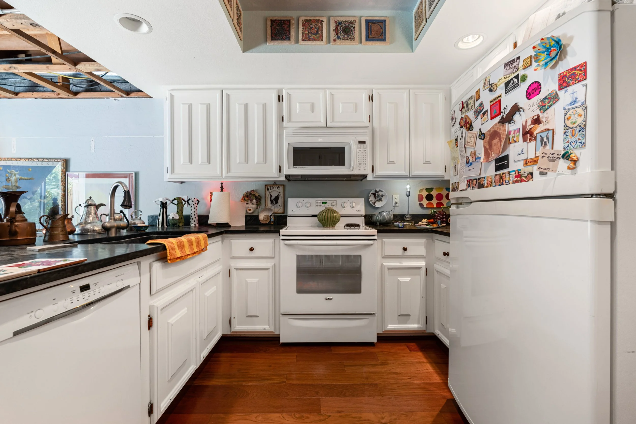 A kitchen with white cabinets, a stove, microwave, and refrigerator covered with colorful magnets and pictures. Wooden floor and various decorative items on the countertops.