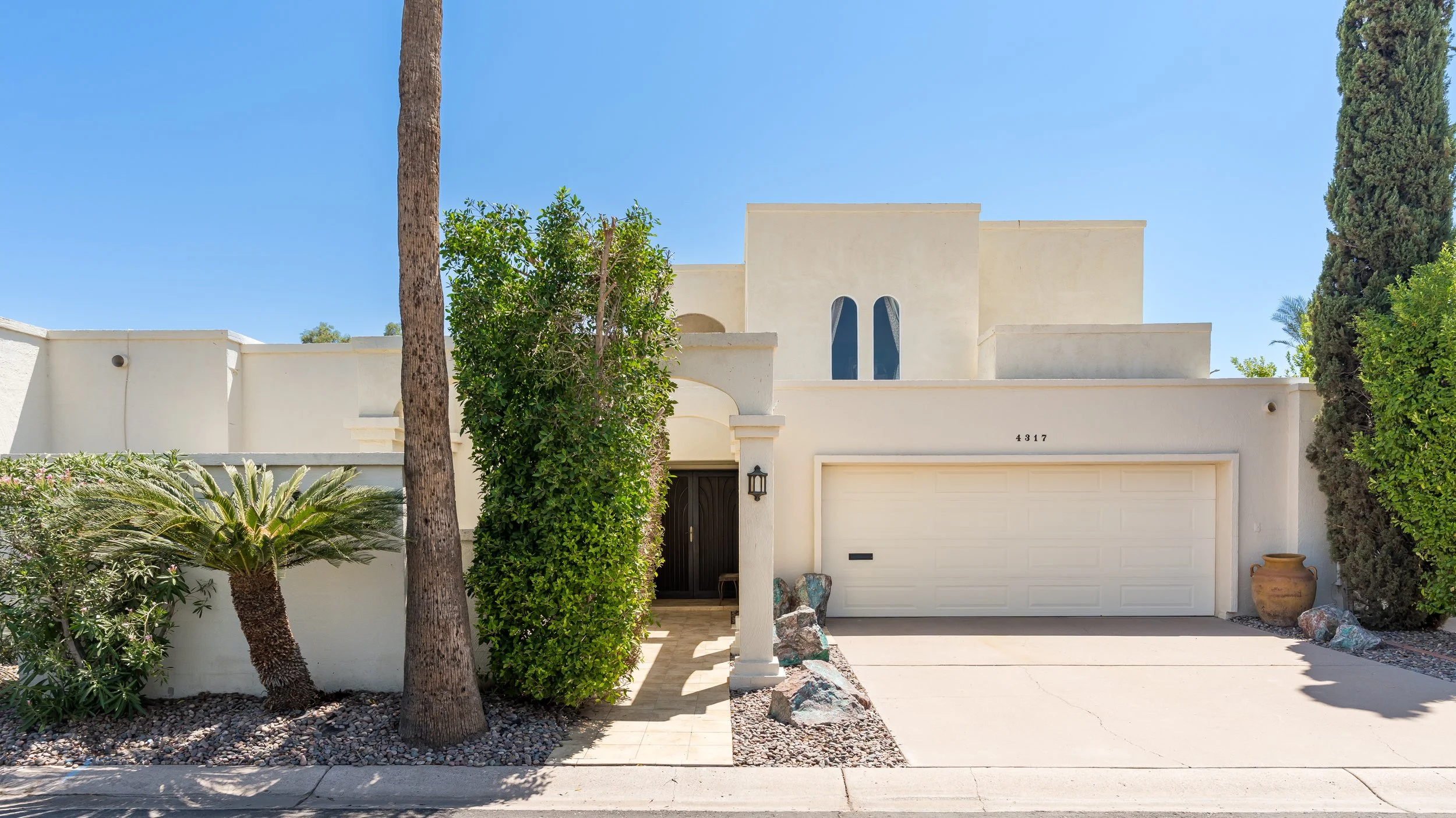 White modern house with a flat roof, a garage door, and an arched front door, surrounded by desert plants, tall trees, and decorative rocks, under a clear blue sky.