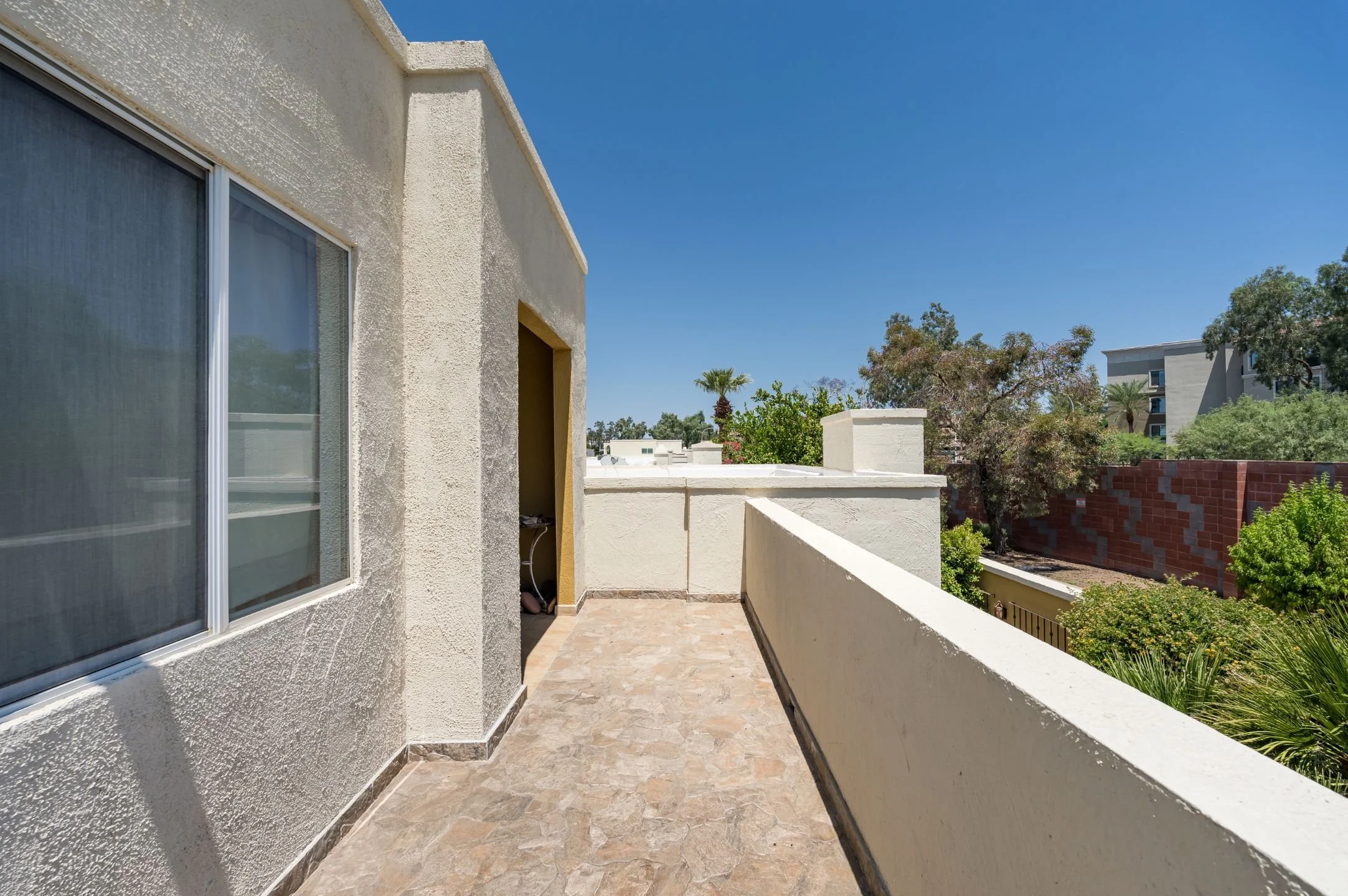 Balcony with beige tiles, white stucco walls, and a glass sliding door, overlooking trees and buildings under a clear blue sky.