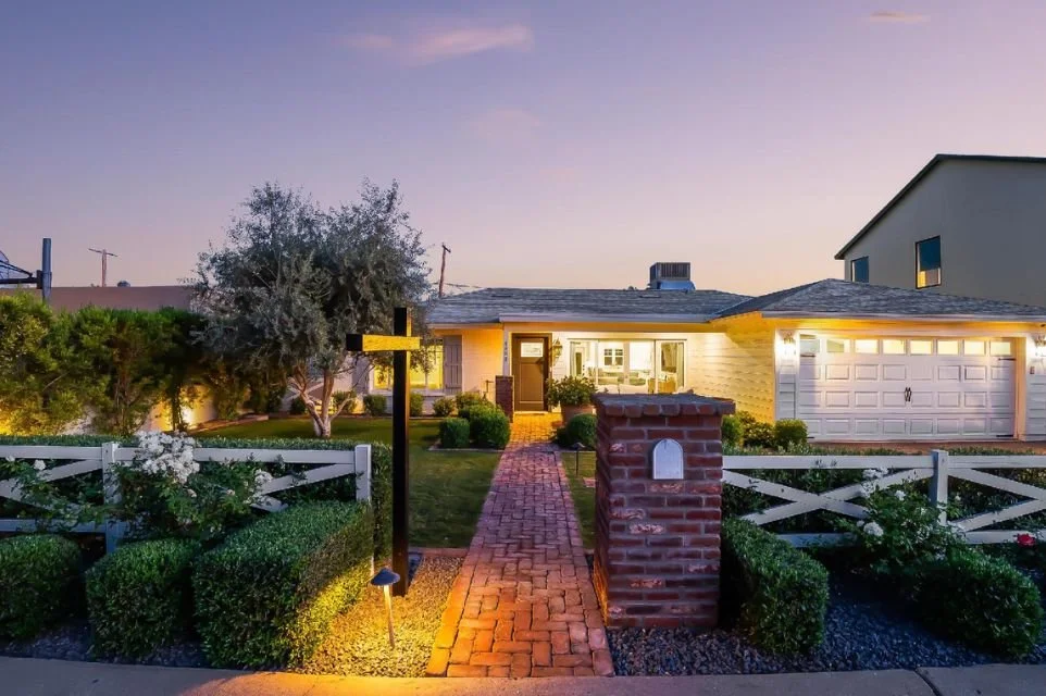 Arcadia Lite home exterior at dusk with illuminated brick walkway, white picket fence, manicured landscaping, and inviting curb appeal in Phoenix Arizona