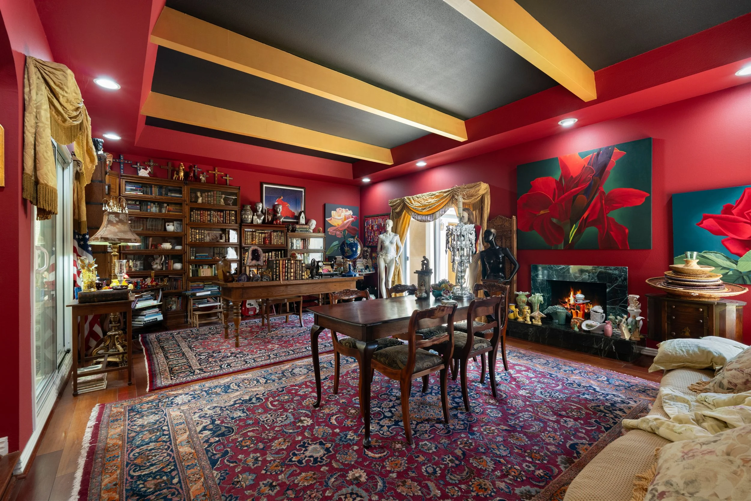 A cozy living room with red walls, a fireplace, a large wooden bookcase filled with books, decorative items, and crosses on top. There are various sculptures and a globe on a table, a Persian rug on the wooden floor, and a dining table with chairs. The room is decorated with large floral paintings, draped curtains, and a variety of decorative objects, creating an eclectic and artistic atmosphere.