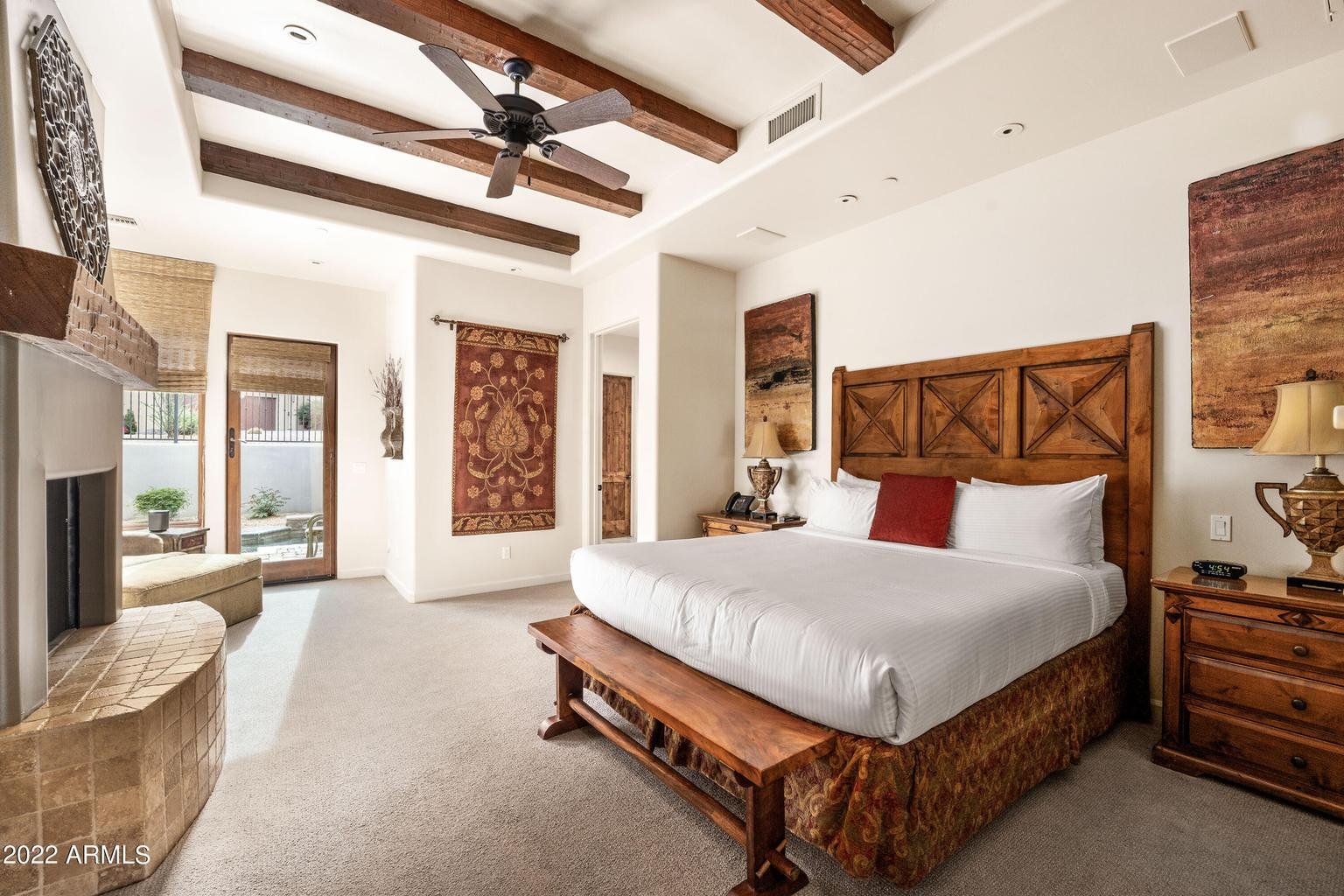 A spacious bedroom with a large wooden bed, white bedding, and red pillows. There are wooden nightstands with lamps, a wall-mounted art piece, and a ceiling fan with wooden beams. The room has a sliding glass door leading to an outdoor area with a pool and patio.