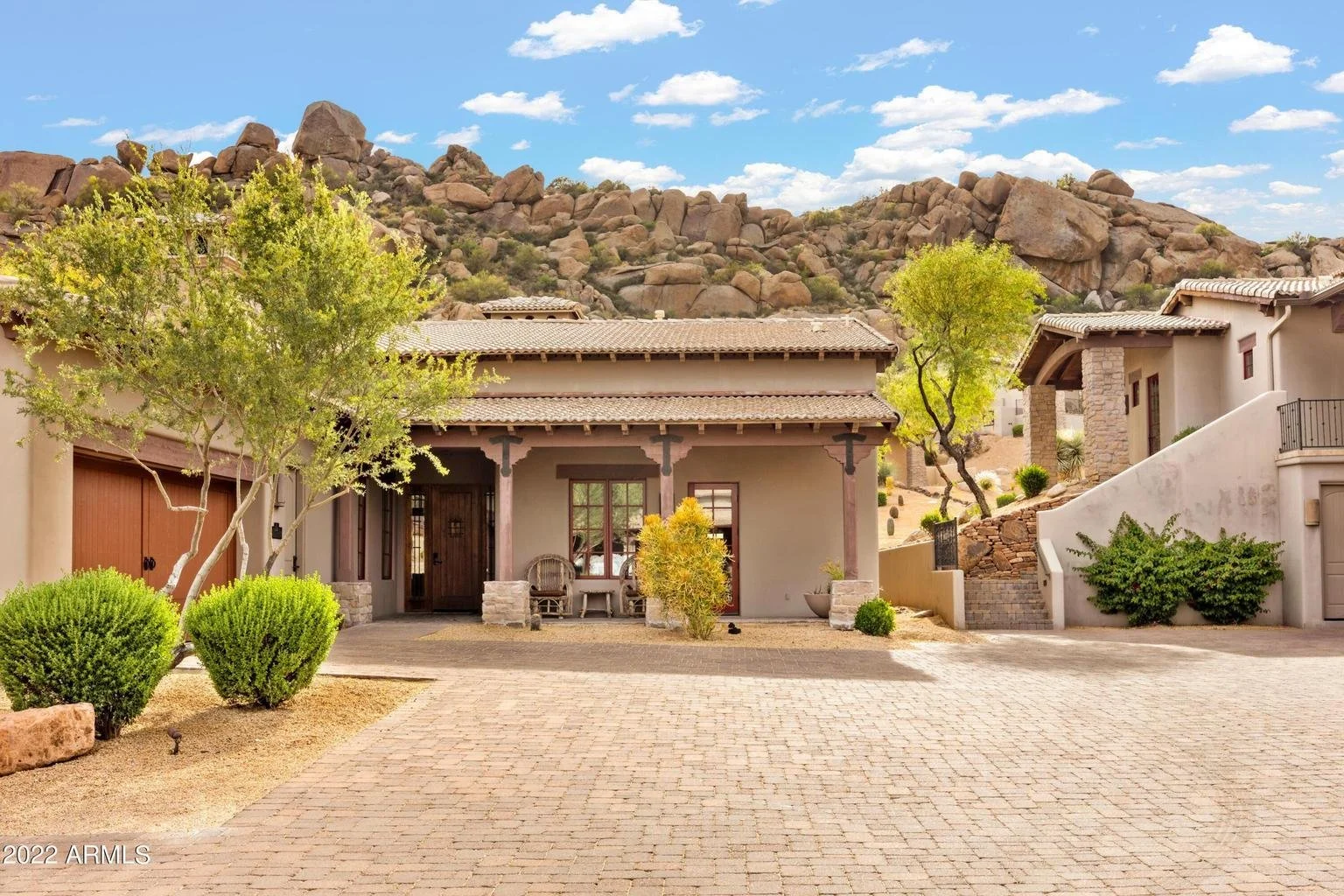 Front view of a southwestern style house with a cobblestone driveway, desert landscaping, and rocky hills in the background under a partly cloudy sky.