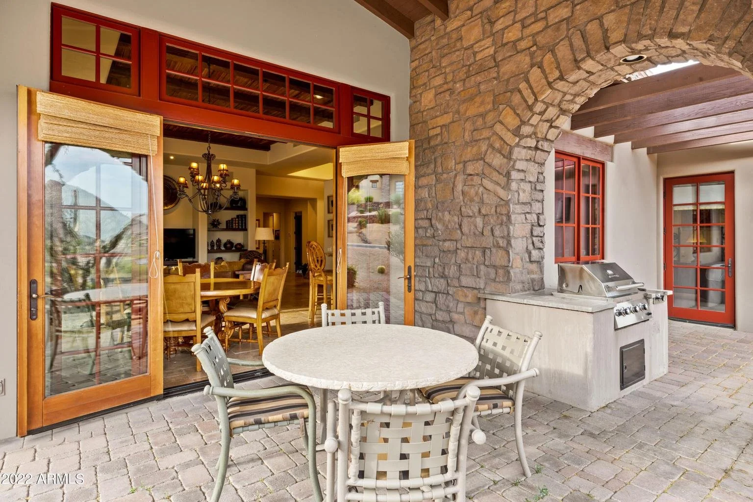 Outdoor patio area with a round table, four chairs, a built-in grill, brick paving, and a stone archway leading to a house with glass sliding doors and red window frames.