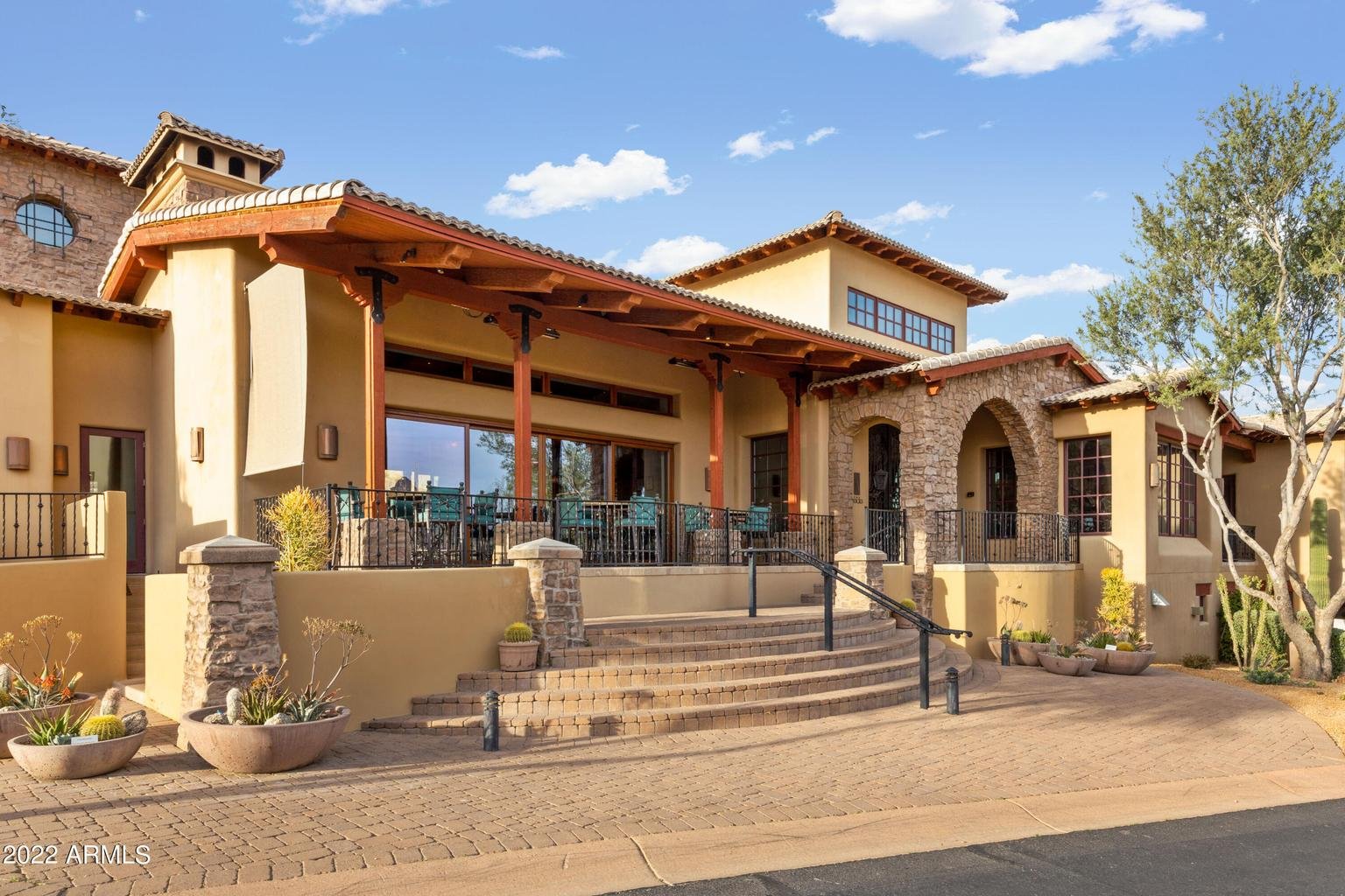 A modern residential house with a stone and stucco exterior, large front steps, and a spacious outdoor patio with railing, in a desert landscape with cacti and trees, under a blue sky with clouds.