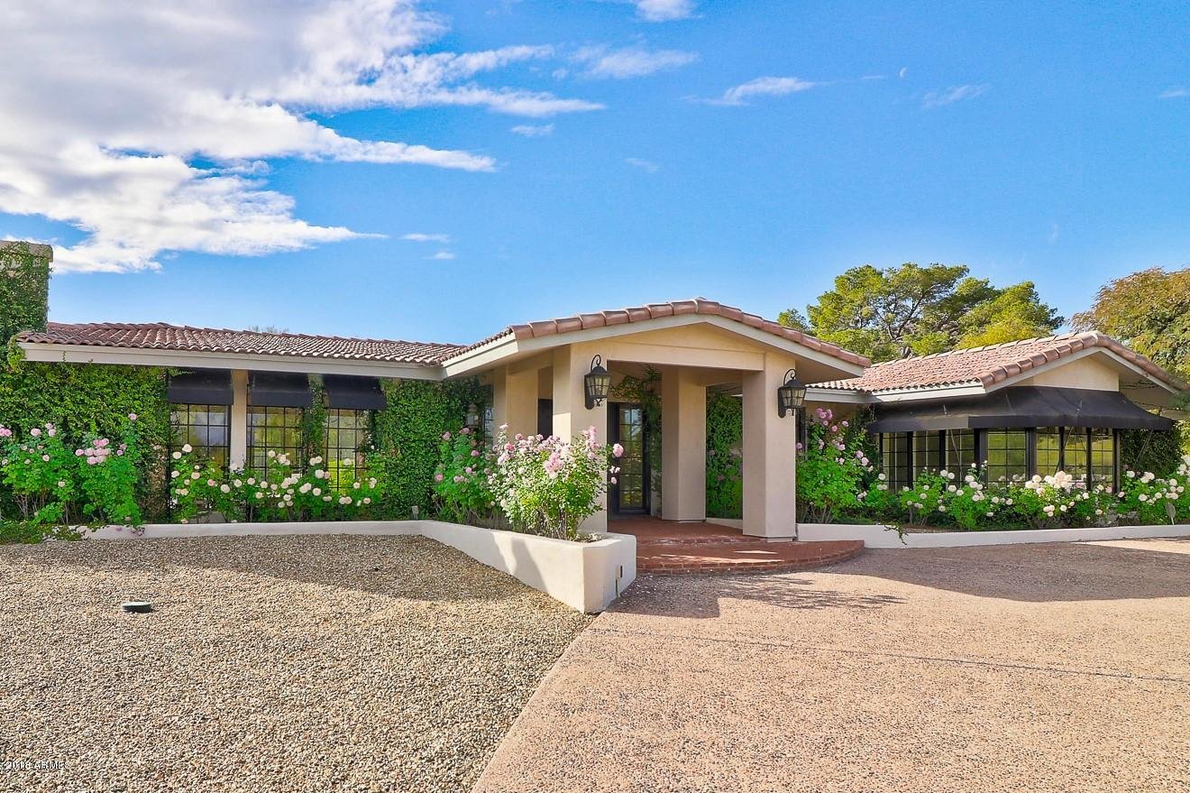 A single-story house with a terracotta tiled roof, surrounded by lush greenery and blooming white and pink flowers, under a bright blue sky with scattered clouds in Paradise Valley