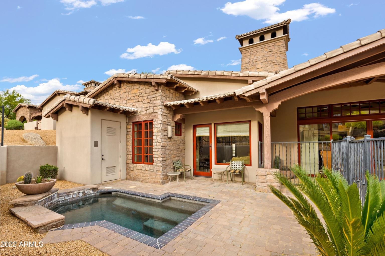 Exterior view of a house with a small pool in the backyard, brick and stone walls, wooden window frames, patio furniture, and desert plants under a blue sky with some clouds.