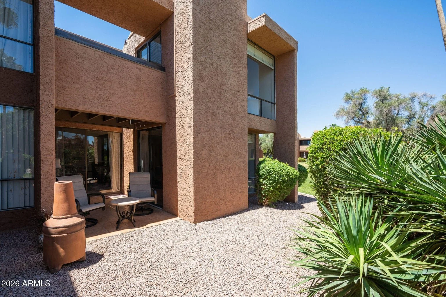 Exterior view of a southwestern-style apartment building with stucco walls, patio area with chairs and small table, surrounded by desert landscaping and bushes under a clear blue sky.