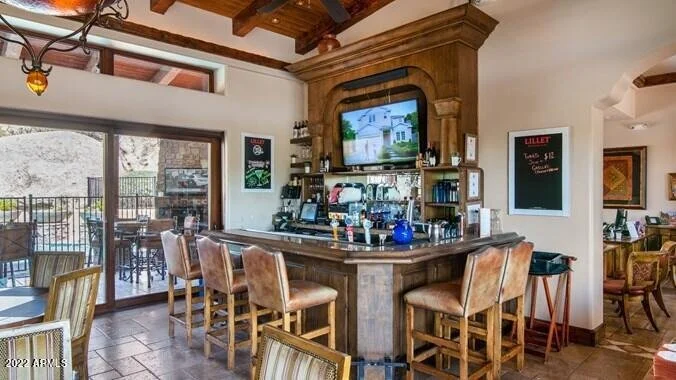Scottsdale community clubhouse bar and lounge featuring wood-beamed ceilings, a large wooden bar with seating, and glass doors opening to an outdoor pool area.