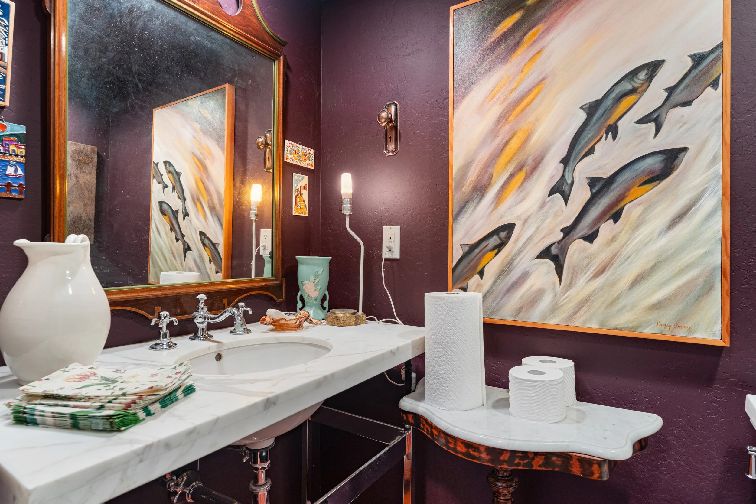 A bathroom countertop with a white sink, silver faucet, colorful folded napkins, and a white vase. Two stacked rolls of toilet paper and a paper towel roll are on a white marble surface. The wall behind is painted dark purple with framed artwork of diving fish. There are two vintage wall lamps and electrical outlets visible.