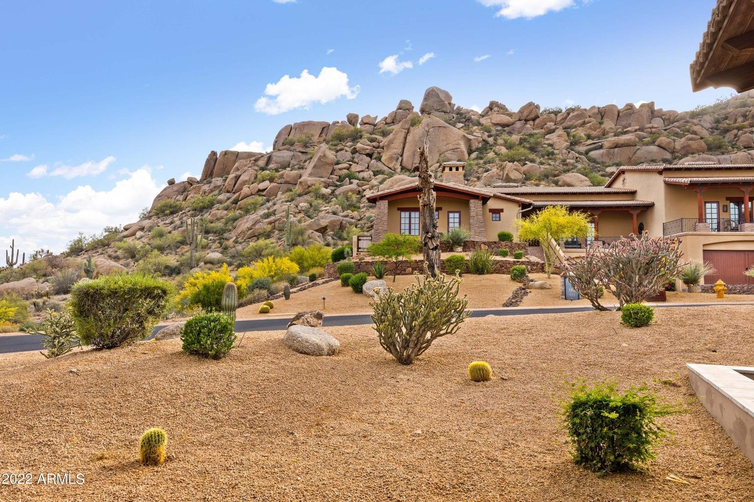 Desert landscape with a house at the foot of rocky hill, with various desert plants including cacti and shrubs, under a partly cloudy blue sky.