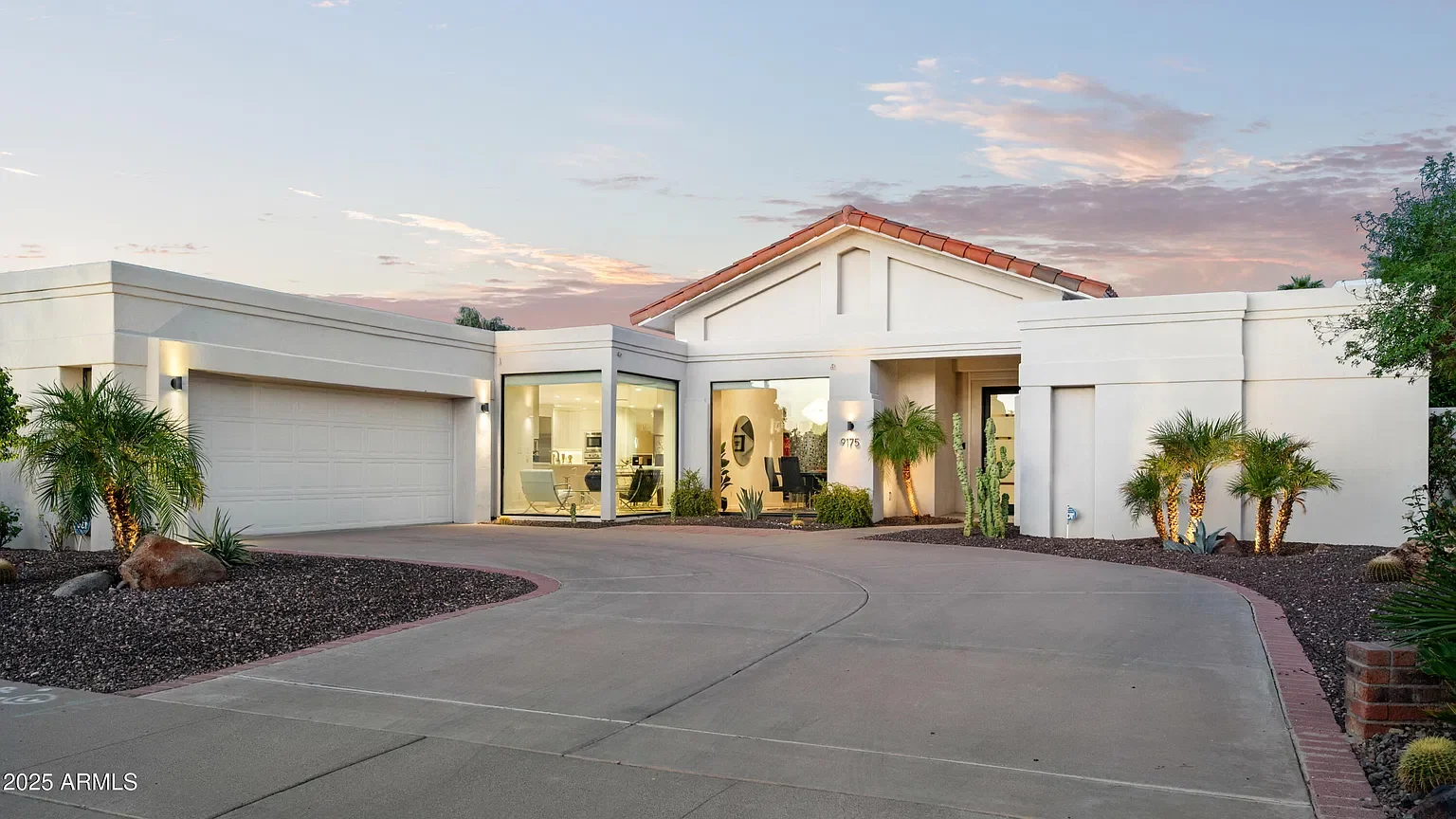 A modern white house with a curved driveway, desert landscaping with rocks, cacti, and small palm trees, and large windows at sunset.