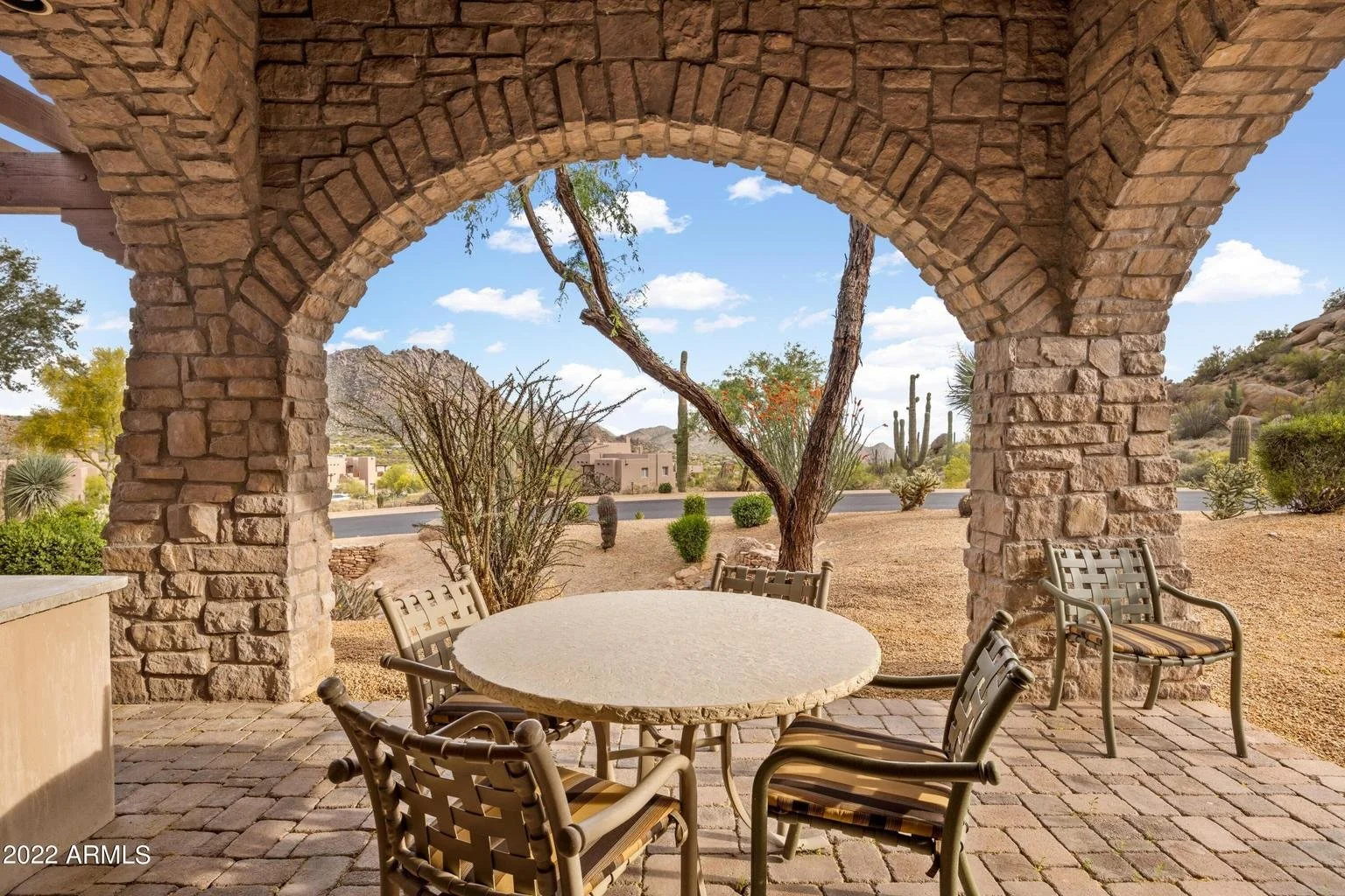 Outdoor patio area with a round table and four chairs under a stone archway, desert landscape with cacti and mountains in the background, clear blue sky with some clouds.