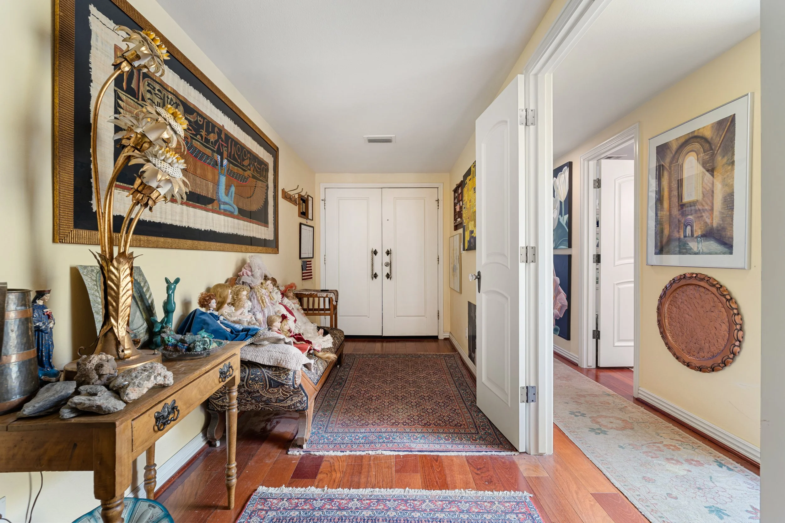 A hallway in a home decorated with artwork on the walls, a wooden console table with decorative items and dolls, and a seating area. The hallway leads to two closed doors, with a door open revealing another room. The flooring is hardwood with various rugs, and the ceiling is plain white.