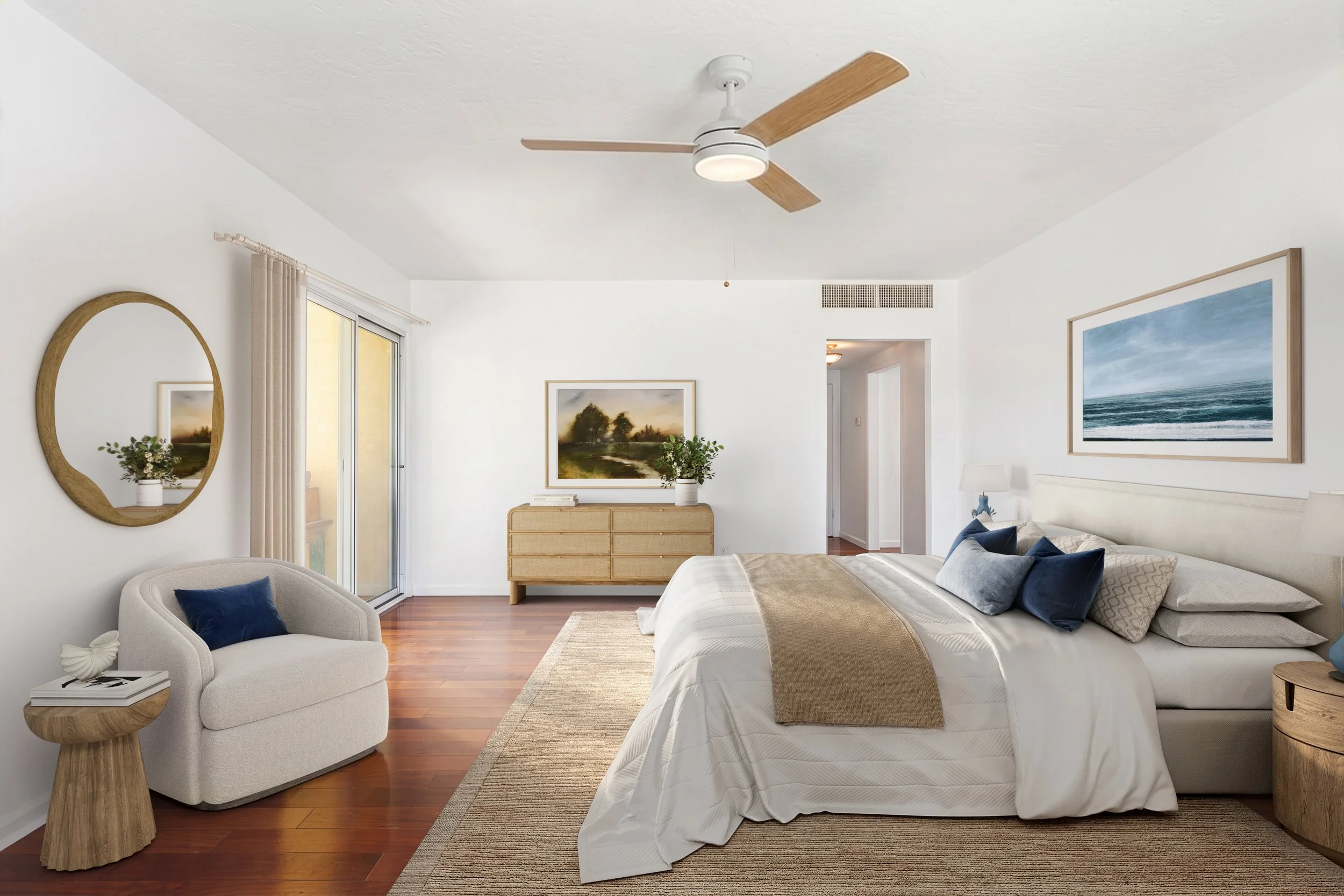 A bedroom with white walls and a white ceiling fan, a sliding glass door with beige curtains, a beige armchair with a blue pillow, and a bed with white and beige bedding. There is a wooden dresser with framed landscape paintings above it, and wall art including a round mirror and a large framed seascape photo. Wood flooring and a beige rug are also visible.