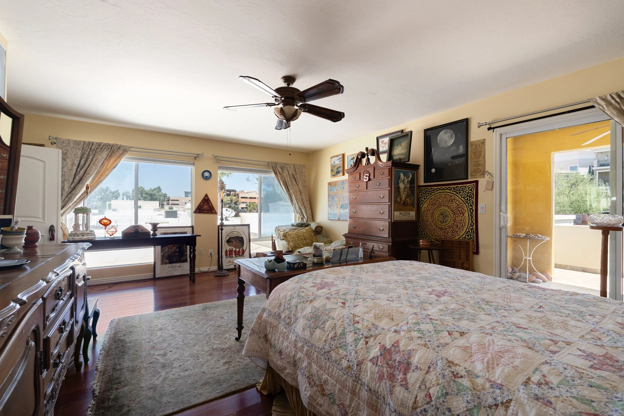 A bedroom with large windows, a ceiling fan, and various framed pictures. There is a bed with a colorful quilt, a wooden dresser, and a sliding glass door leading to a balcony or patio area.