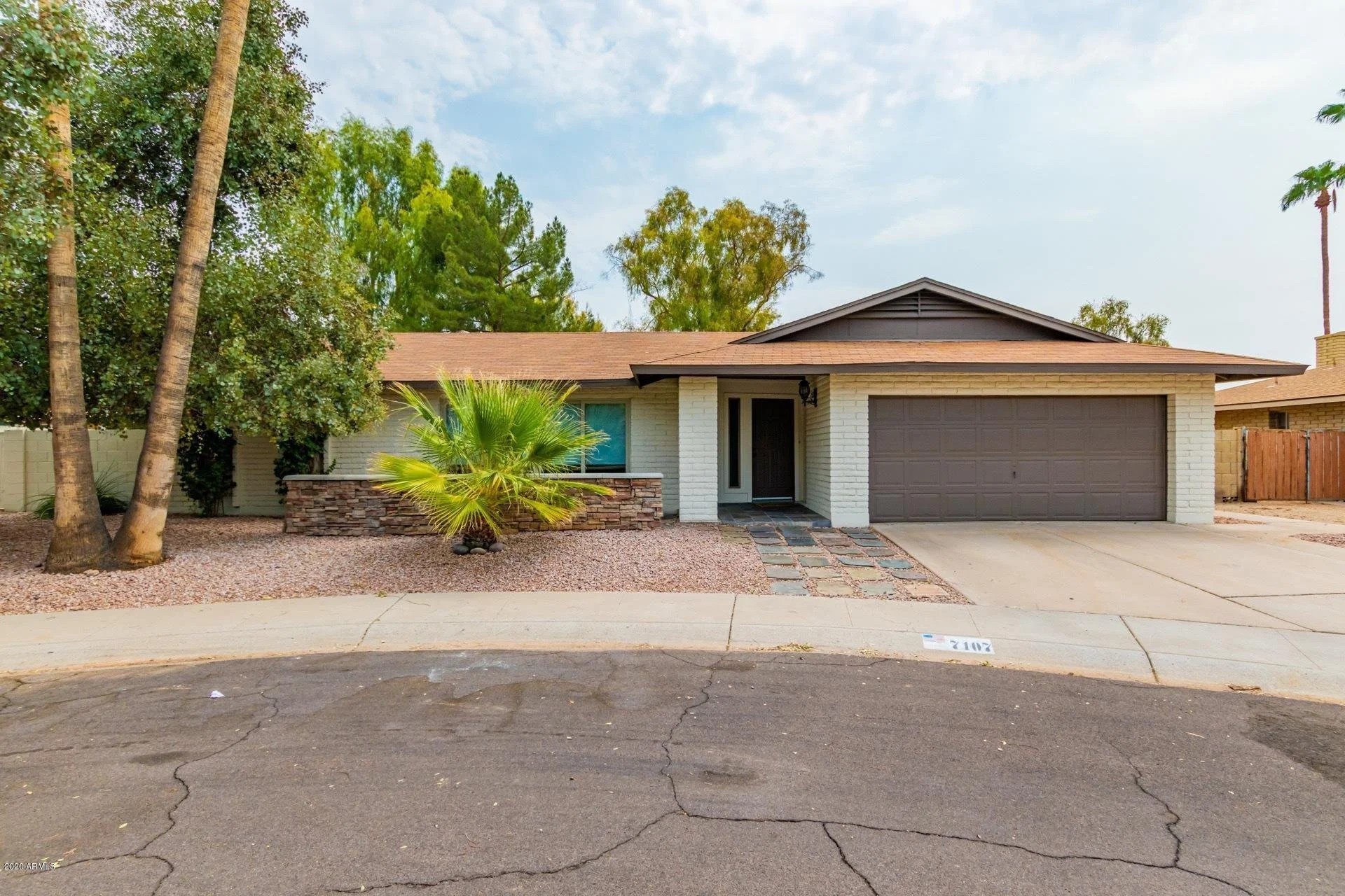 Front view of a single-story house with a brick exterior, gray garage door, and a small front porch with stone stepping stones. There are tall palm and other trees on the left side of the house, and a gravel yard with desert landscaping. The house has a light-colored fence and a sidewalk in front with the house number on the street.