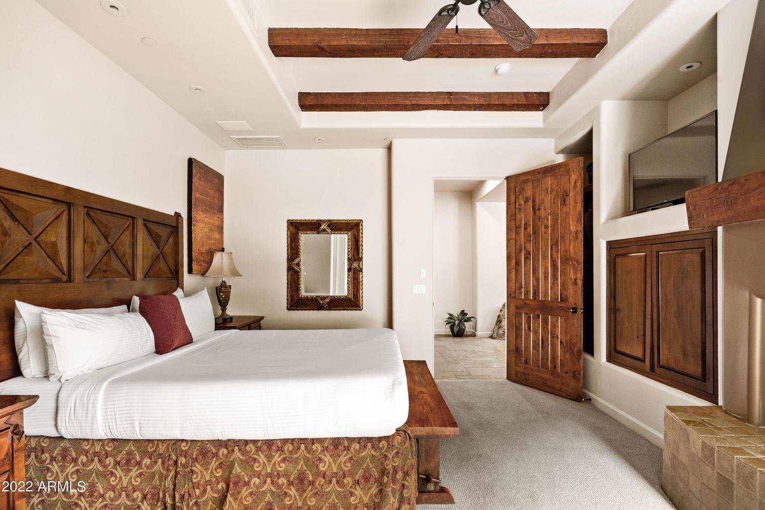 A bedroom with a large bed, wooden headboard, plush pillows, and a nightstand with a lamp. There are wooden beams on the ceiling, a wall mirror, and an open wooden door leading to a room with a potted plant. A TV is mounted above a built-in wooden cabinet.