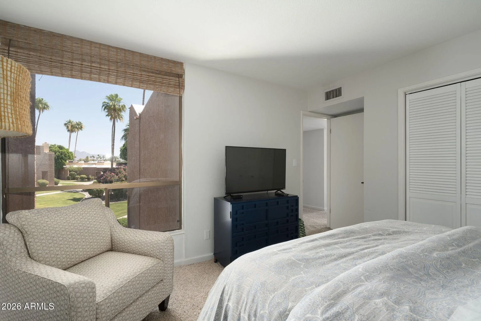 Bedroom at 7401 N Scottsdale Rd #26 in The Shores at McCormick Ranch, Scottsdale, featuring neutral carpeting, a wooden bed frame, a large window with palm tree views, and a spacious closet.
