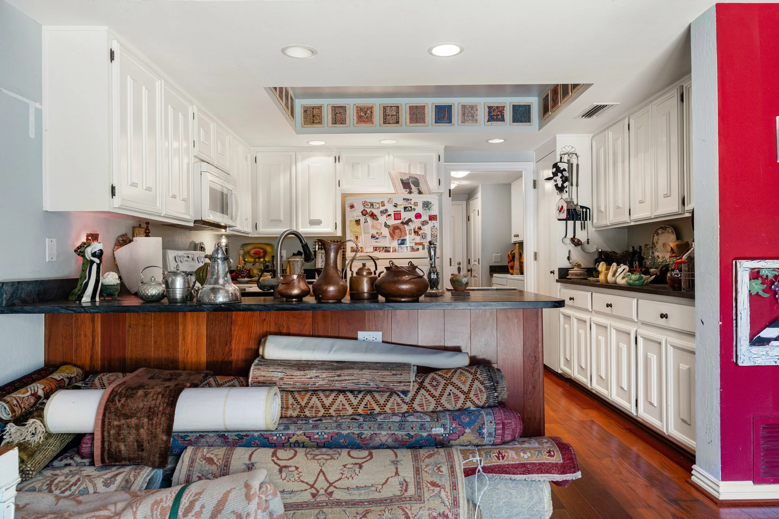 A kitchen with white cabinets, a black countertop, and a variety of decorative items including vases and figurines. A red wall is partially visible on the right side, and a stack of rolled up rugs is on the floor in the foreground.