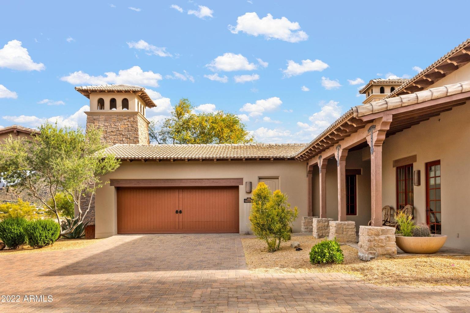 Front view of a house with a desert landscape, including a paved driveway, southwestern architectural style, and blue sky with scattered clouds.