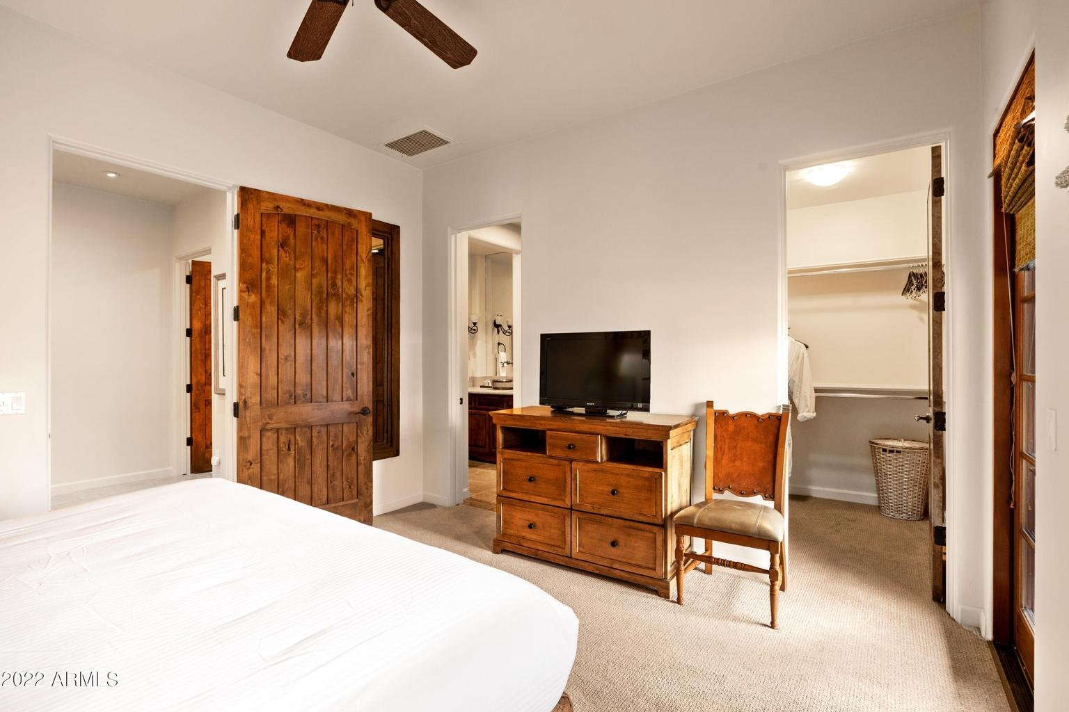 A bedroom with a bed on the left, a wooden dresser with a small TV on top, an empty chair next to the dresser, and two open closet doors showing clothes and a laundry basket. Wooden door and window shutters are also visible.