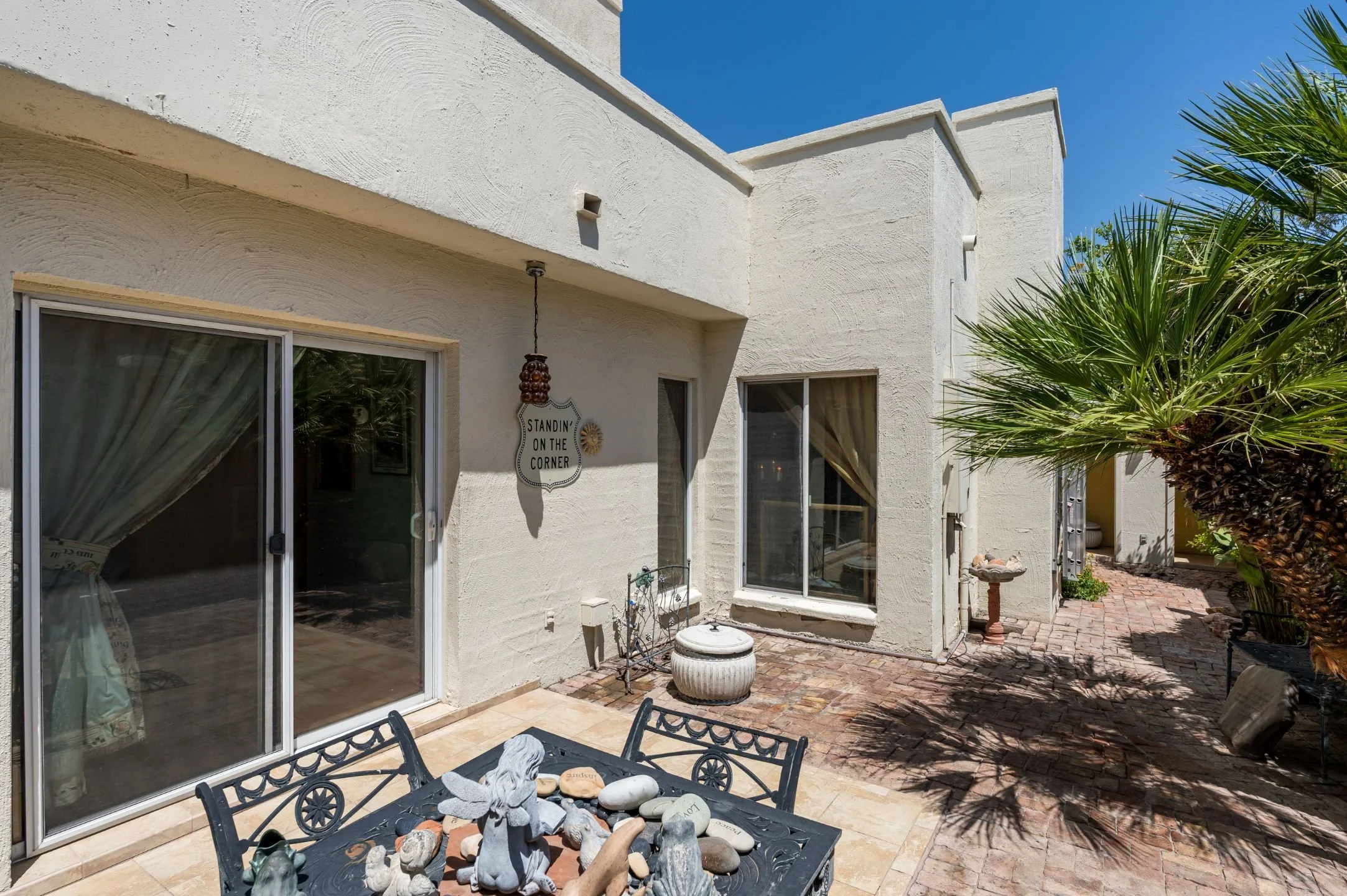 A backyard patio with a black metal table and chairs, a decorative water fountain, a white stucco house wall with sliding glass doors, a sign that reads 'Standing on the corner,' and lush green palm trees under a bright blue sky.