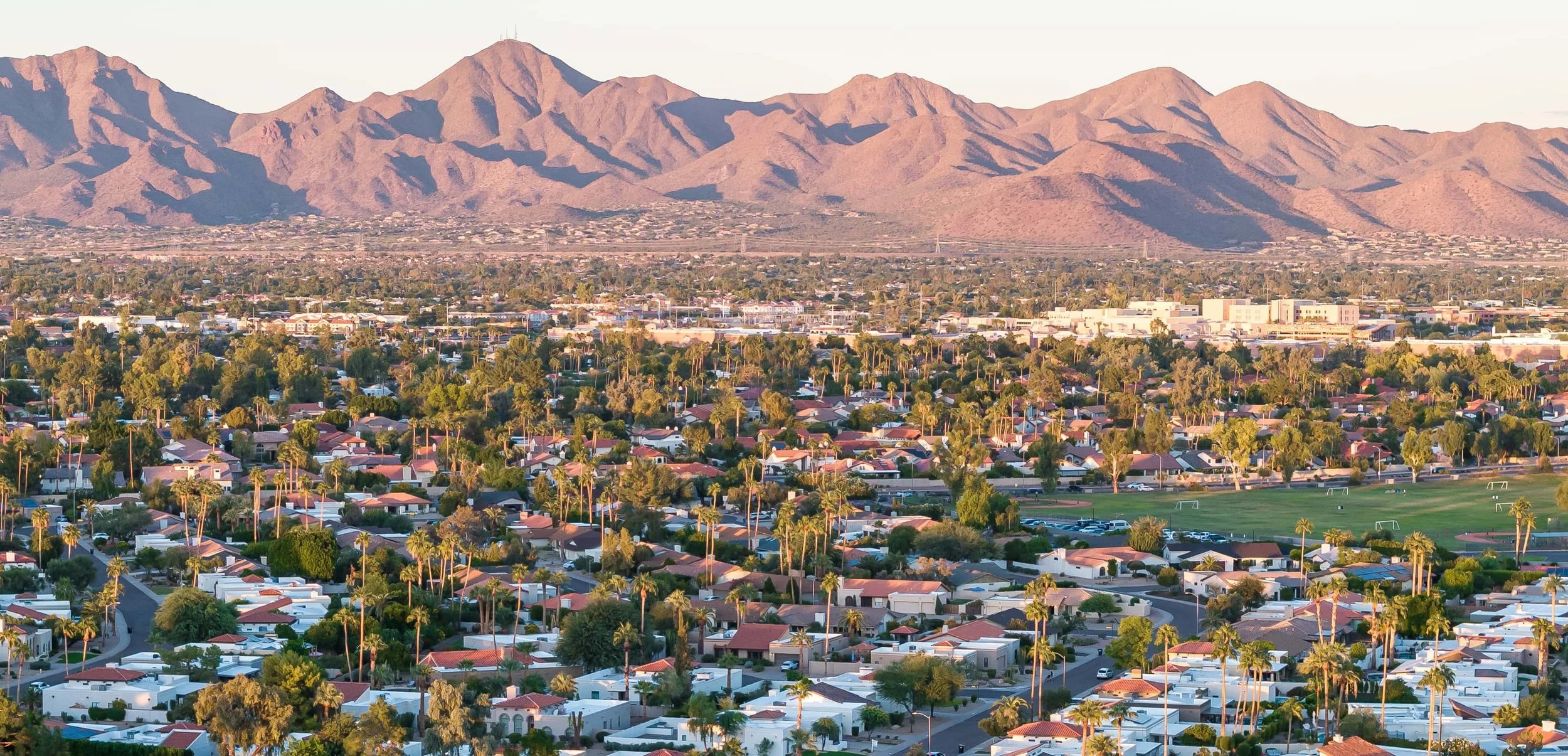 Aerial view of a suburban neighborhood with houses, trees, and a park, with mountains in the background during sunset.