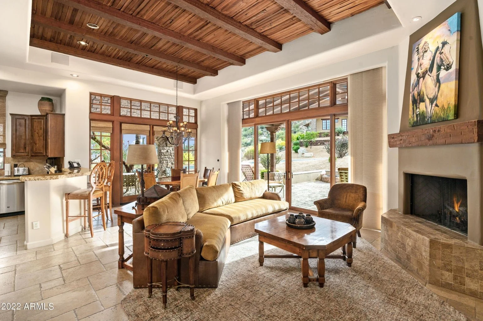 Luxury Scottsdale home living room featuring exposed wood-beamed ceilings, a stone-tiled fireplace, large sliding glass doors, and views of the desert landscape.