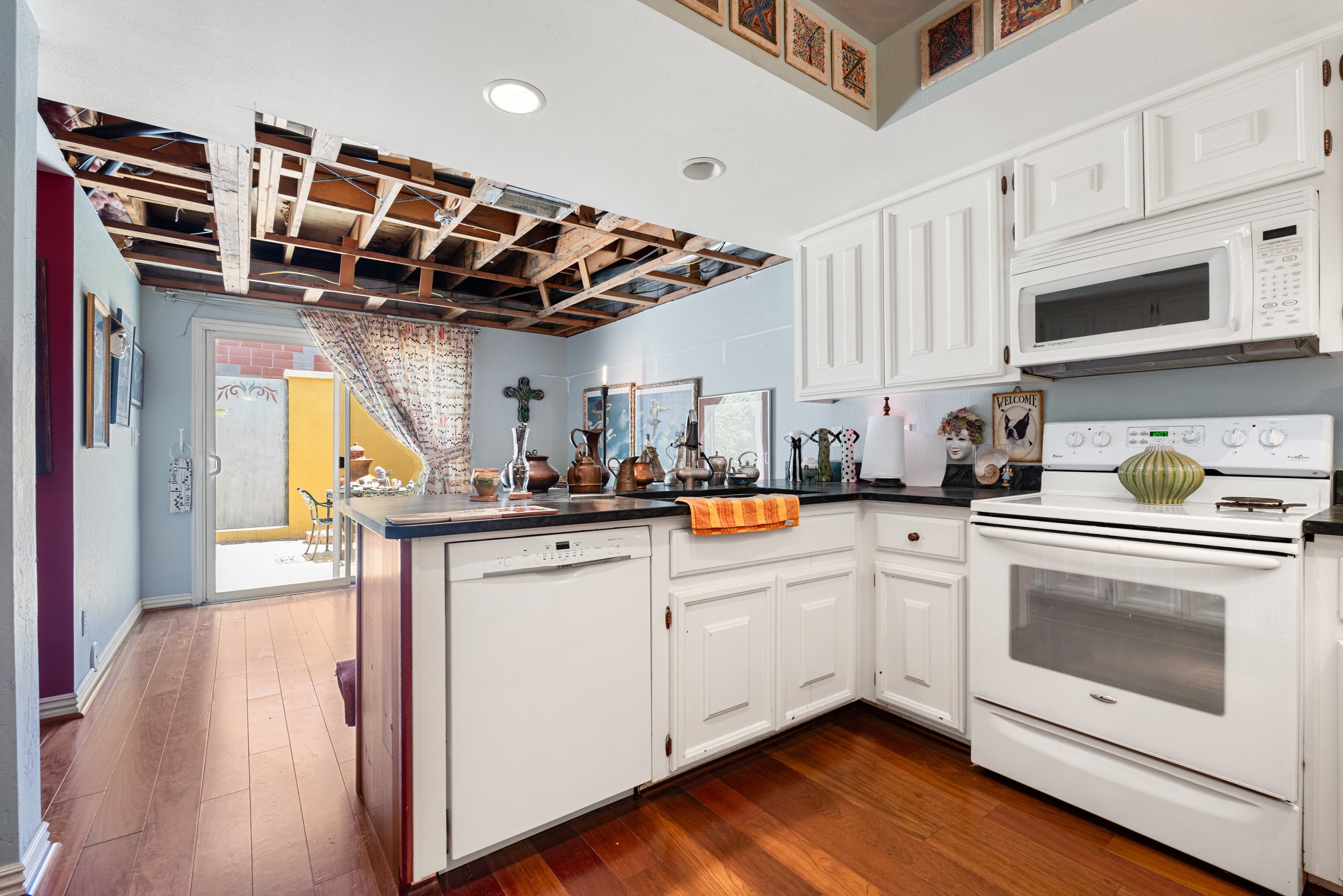 A kitchen area with white cabinets, a microwave oven, a stove, and a countertop with various decorative items. The ceiling has an unfinished section with exposed wooden beams and insulation. There is a sliding glass door leading to an outdoor patio, visible through a partially open curtain.