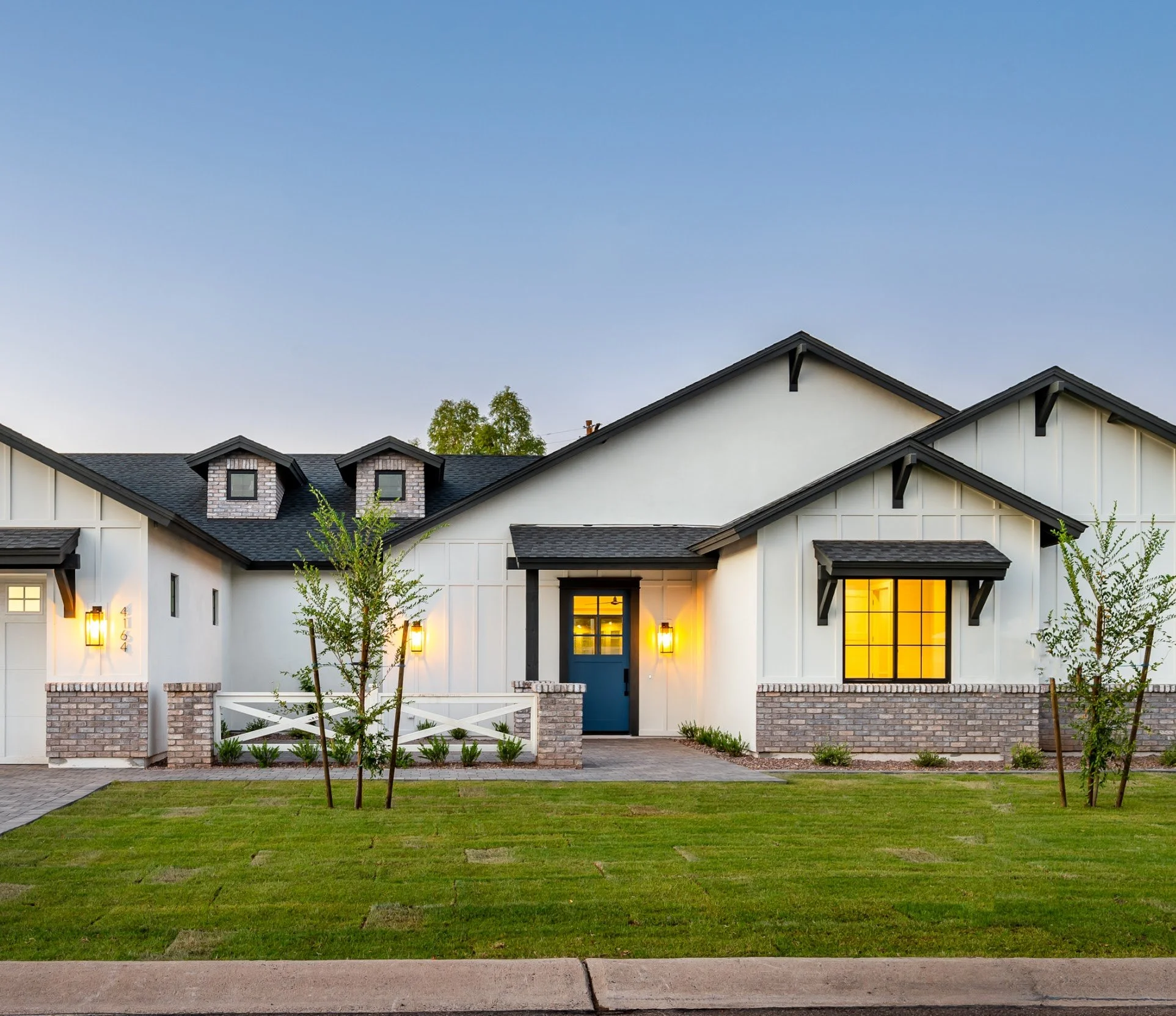 Modern suburban house with white siding, brick accents, black roof, and warm yellow interior lights visible through large windows, front yard with green grass and small trees, driveway on the left, evening sky in the background.