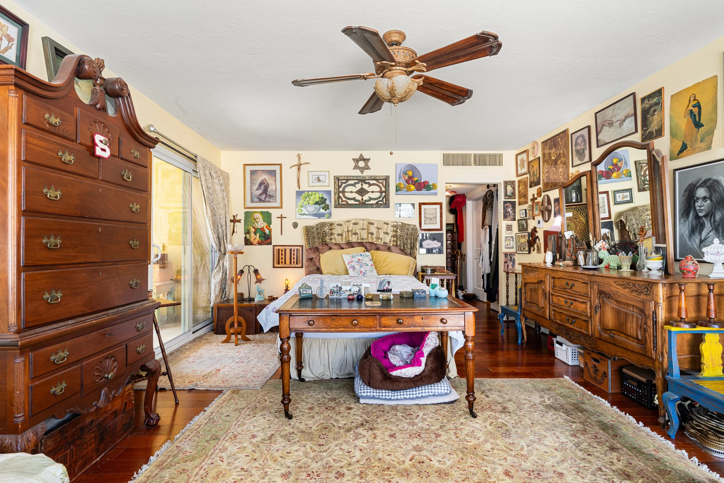 A cozy vintage-style bedroom decorated with numerous religious and artistic artworks, a large wooden dresser on the left, a bed with pillows in the center, and a wooden cabinet on the right. The room has a ceiling fan, hardwood floors, and a rug.