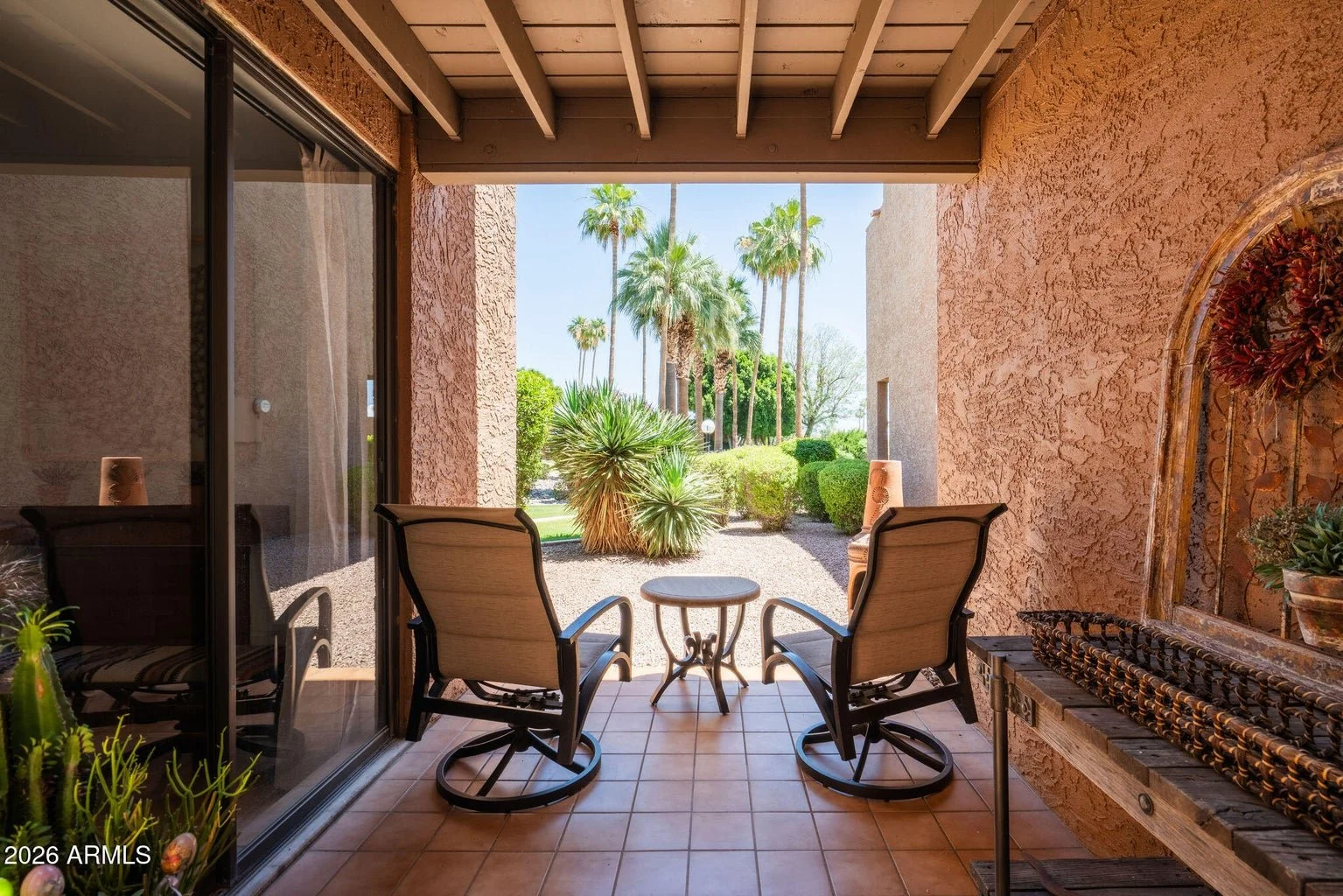 Covered outdoor patio at 7401 N Scottsdale Rd #26 in The Shores at McCormick Ranch, featuring two lounge chairs, a small table, tiled flooring, exposed ceiling beams, and views of palm trees and landscaped grounds.