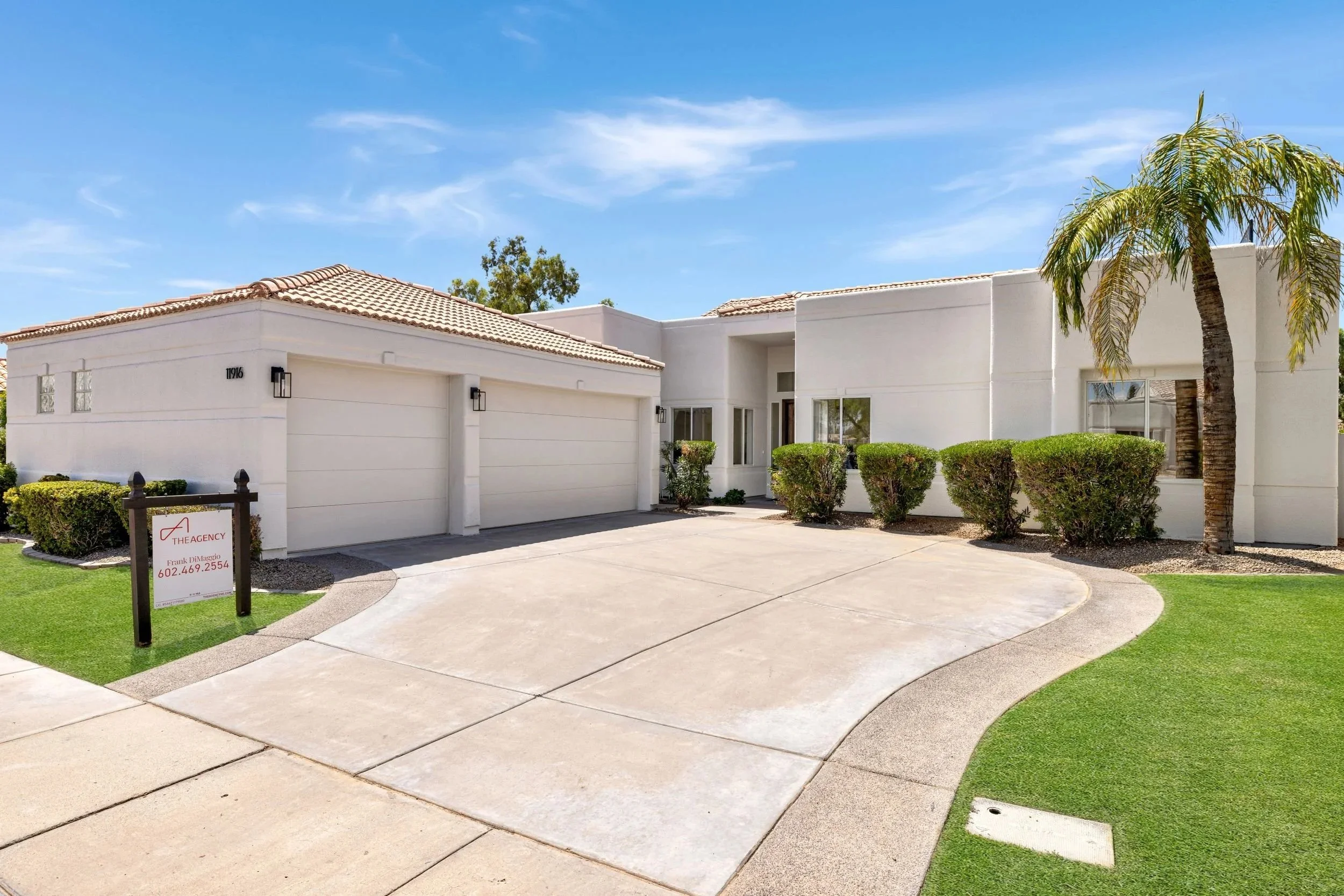 A white modern house with a driveway, green bushes, and a palm tree under a blue sky.