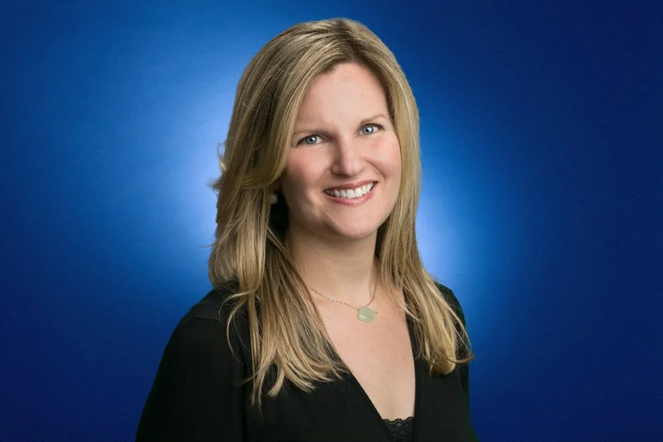 A woman with long blonde hair wearing a black top and a light green necklace, smiling against a blue background.