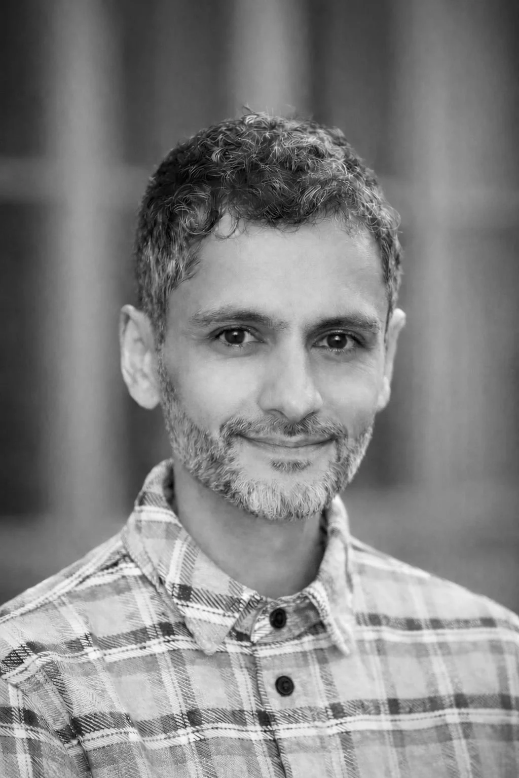 Black and white portrait of a man with short, curly hair and a beard, wearing a plaid shirt, smiling subtly at the camera.