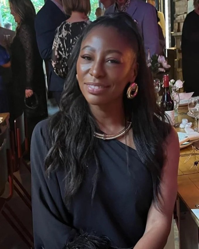 A woman with long, wavy black hair, wearing large gold hoop earrings and a silver necklace, is smiling at the camera in a social setting with people and a dining table in the background.