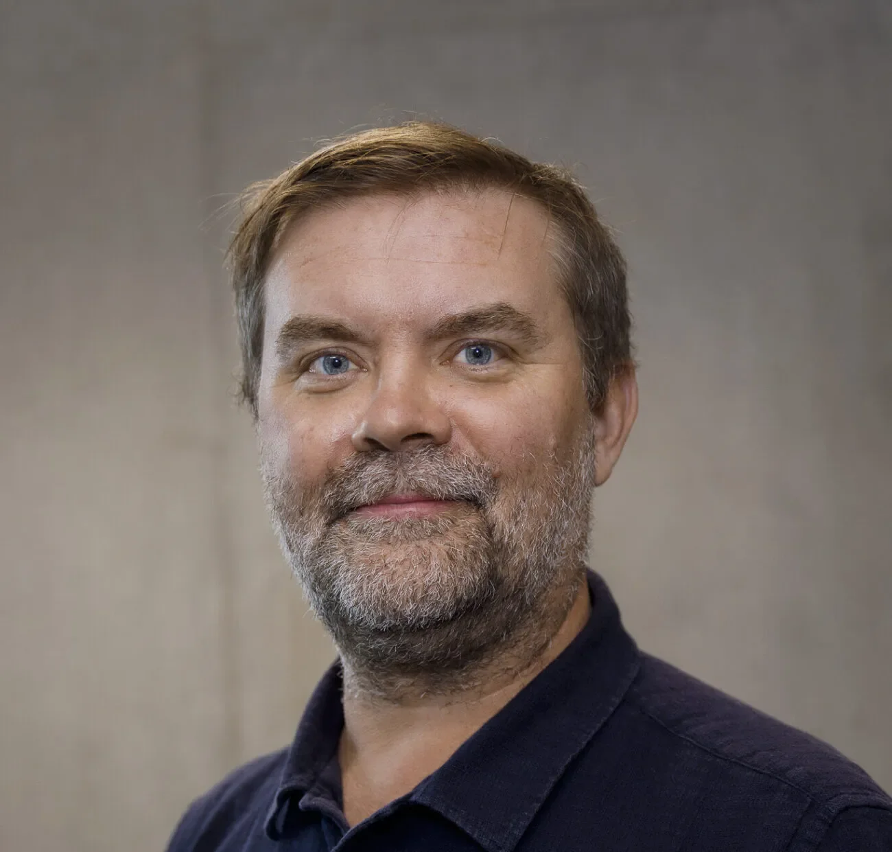 Close-up portrait of a middle-aged man with short brown hair, blue eyes, a beard, and a mustache, wearing a dark navy shirt, against a plain neutral background.