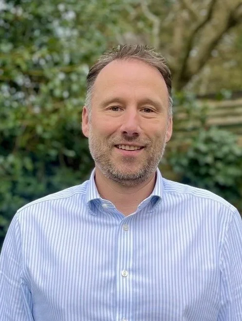 A smiling man with light skin, short brown hair, and a beard, wearing a light blue striped button-up shirt, standing outdoors with greenery and trees in the background.